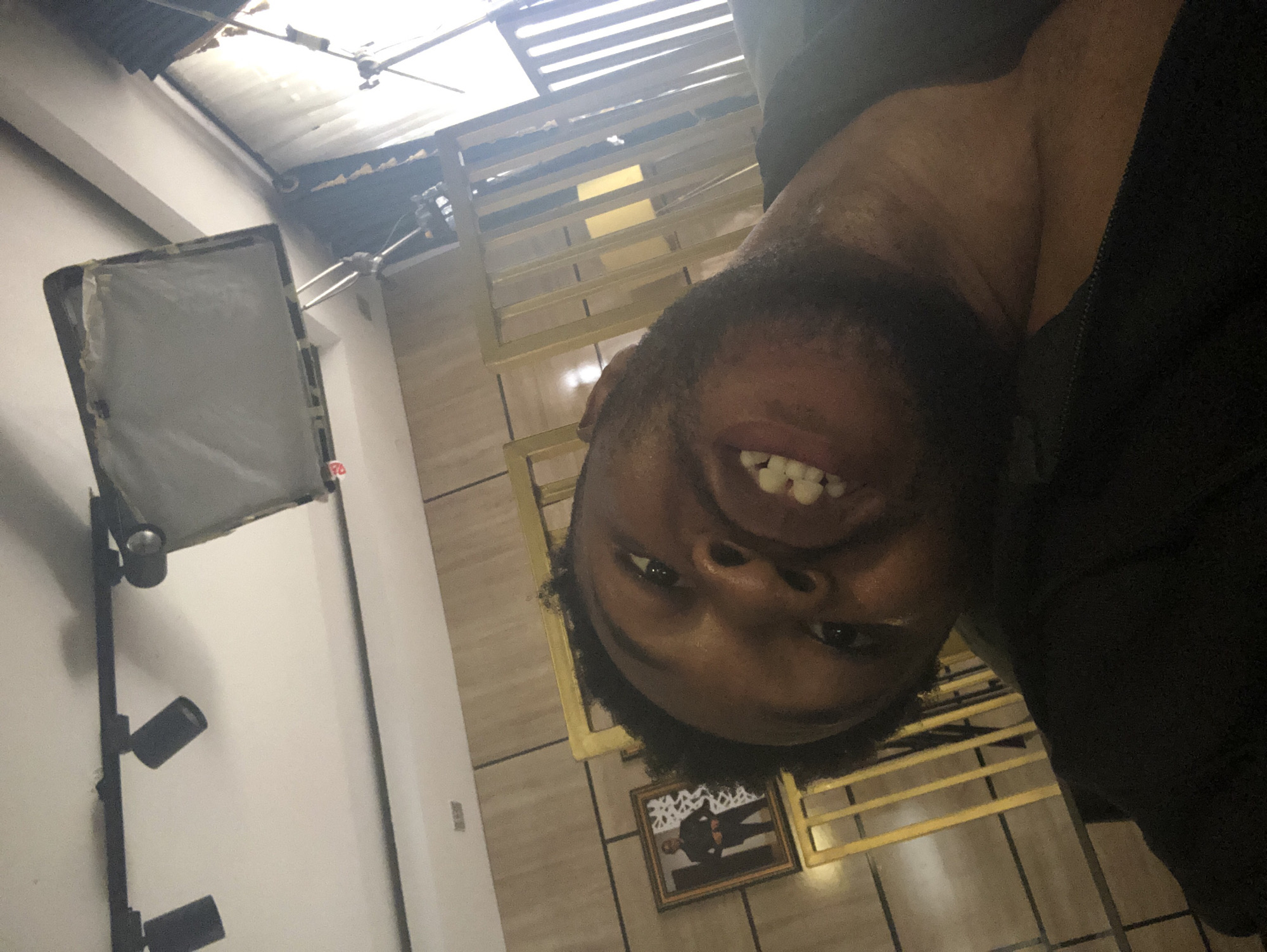 The image is rotated 90 degrees counter-clockwise, showing a close-up, upside-down portrait of a smiling person indoors.
The primary subject is a person with dark skin, short dark hair, and a beard, looking directly at the camera with a wide smile, revealing teeth. They appear to be wearing a dark-colored shirt or jacket, visible on their chest and shoulder.
The setting is an indoor space, likely a studio or commercial area, characterized by varied architectural elements. To the left (when viewed upright), a rectangular softbox light diffuser is mounted on a stand, accompanied by several smaller, dark-colored spot lights. Further in the background, a white wall is visible.
To the right of the person, a gold-colored metal railing or banister is prominent. Below this railing, on a tiled floor, rests a framed picture depicting a person in a suit, though details are indistinct. The ceiling features corrugated metal roofing and exposed beams or structural elements, with bright light filtering through some sections.
The overall activity suggests a selfie or a staged photograph, given the close-up, direct gaze, and prominent lighting equipment. The person's smile conveys a positive or cheerful emotion. The artificial lighting dominates the scene, making the time of day unidentifiable. No clearly legible text is visible in the image.