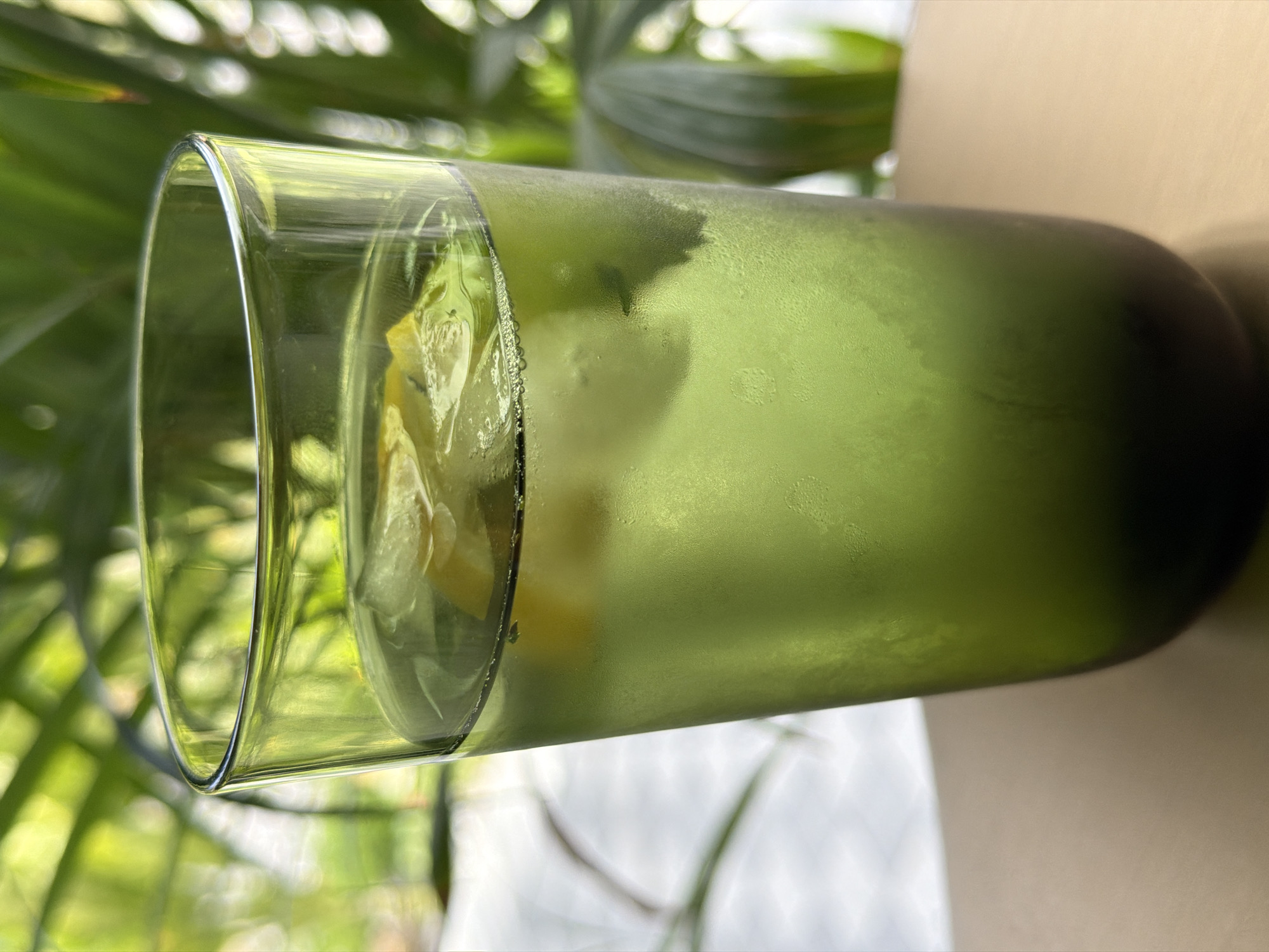 A close-up shot shows a tall, green, frosted glass filled with a light green beverage and ice. Slices of lemon are visible floating in the drink. The glass is condensation-covered, suggesting it is cold. The background is blurred, featuring lush green foliage, consistent with the tropical environment of Ko Samui District, Thailand. The glass sits on a light brown, smooth surface, possibly a table.