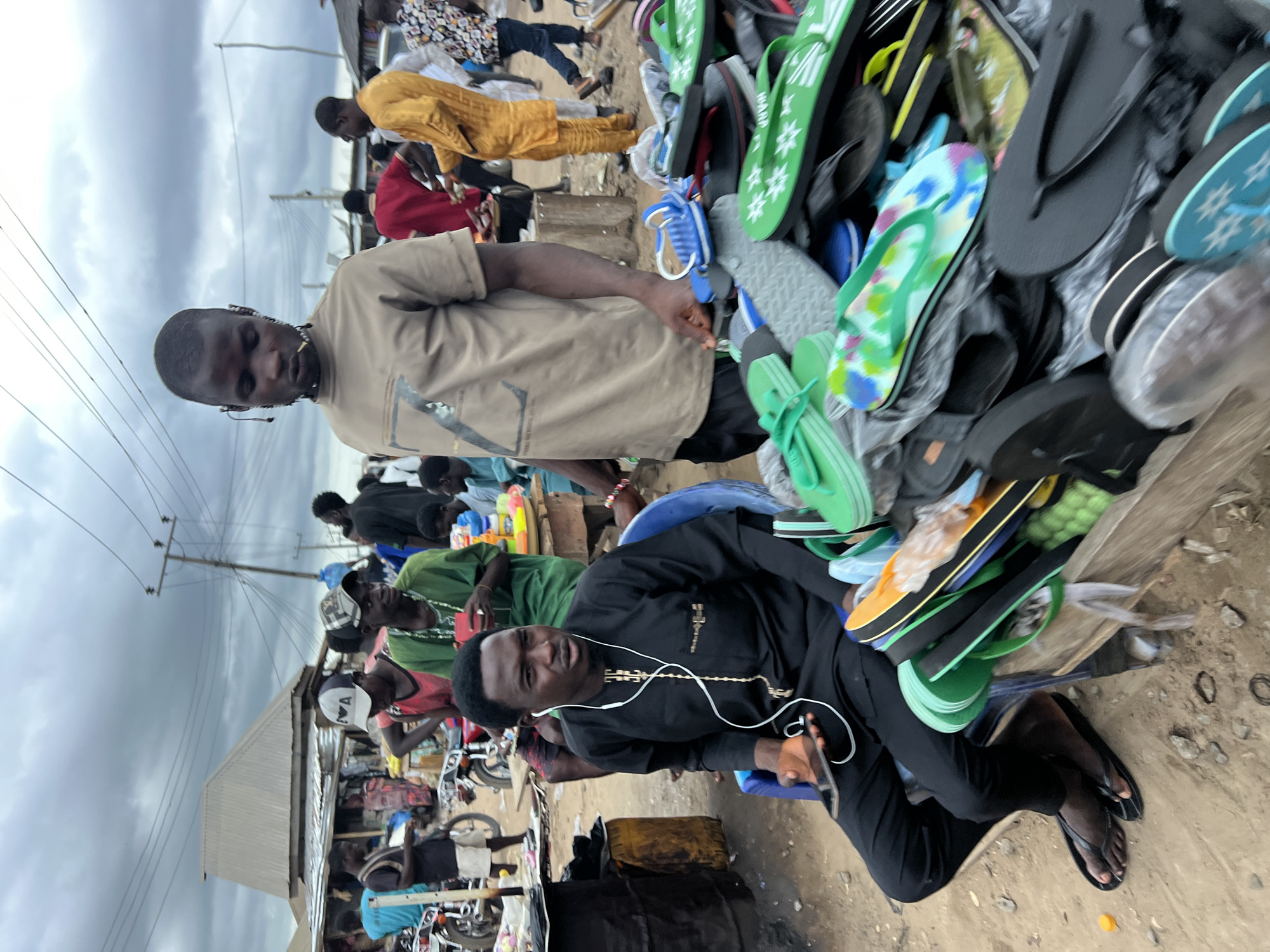 In Oko Amakom, Nigeria, under an overcast sky, a street market scene unfolds. Two men are the primary subjects. In the foreground, seated on a blue plastic chair, a man in a black traditional outfit with white earphones is holding a smartphone and looking to his right. Beside him, a large display of colorful flip-flops and sandals is arranged on a wooden structure. Behind him, standing and looking forward, is another man wearing a tan t-shirt. A bustling market atmosphere is suggested by the presence of other individuals in the background, motorcycles, and makeshift stalls with corrugated metal roofs. The ground is dirt and scattered with small stones.