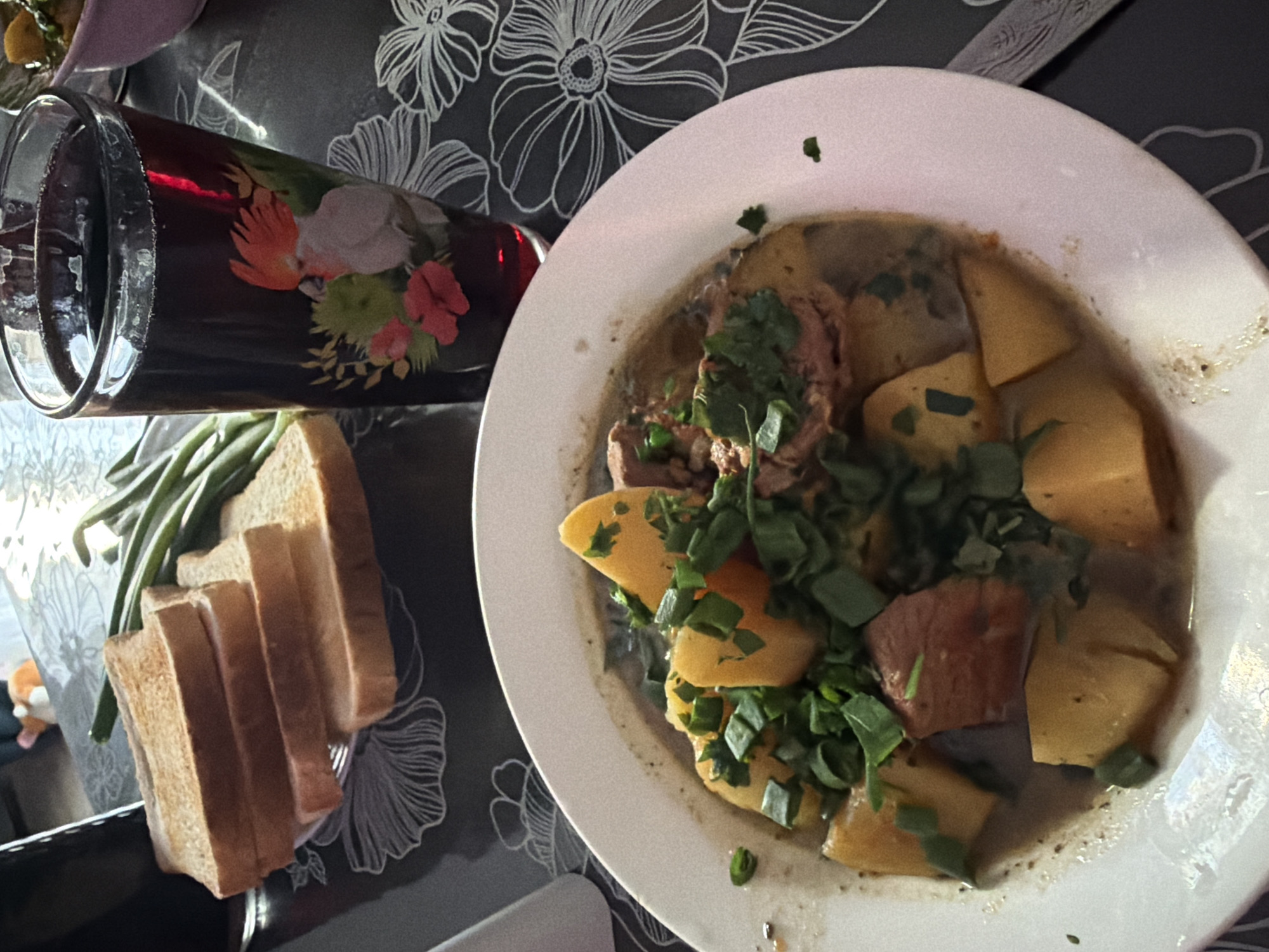 A high-angle, close-up shot shows a table setting with a meal and a drink. In the foreground, a white plate holds a stew with large chunks of meat and potatoes, garnished with chopped green herbs. To the left of the plate, several slices of toasted bread are stacked, next to a bunch of green beans. A tall glass with a dark red beverage, possibly wine, is positioned behind the food, decorated with a colorful bird and floral design. The table appears to be covered with a dark tablecloth featuring a floral pattern. The setting suggests a meal is being served or enjoyed. The lighting indicates it could be daytime or an indoor setting with artificial light. There is no visible text in the image. The location is identified as Krasnyi Krym, Russia.