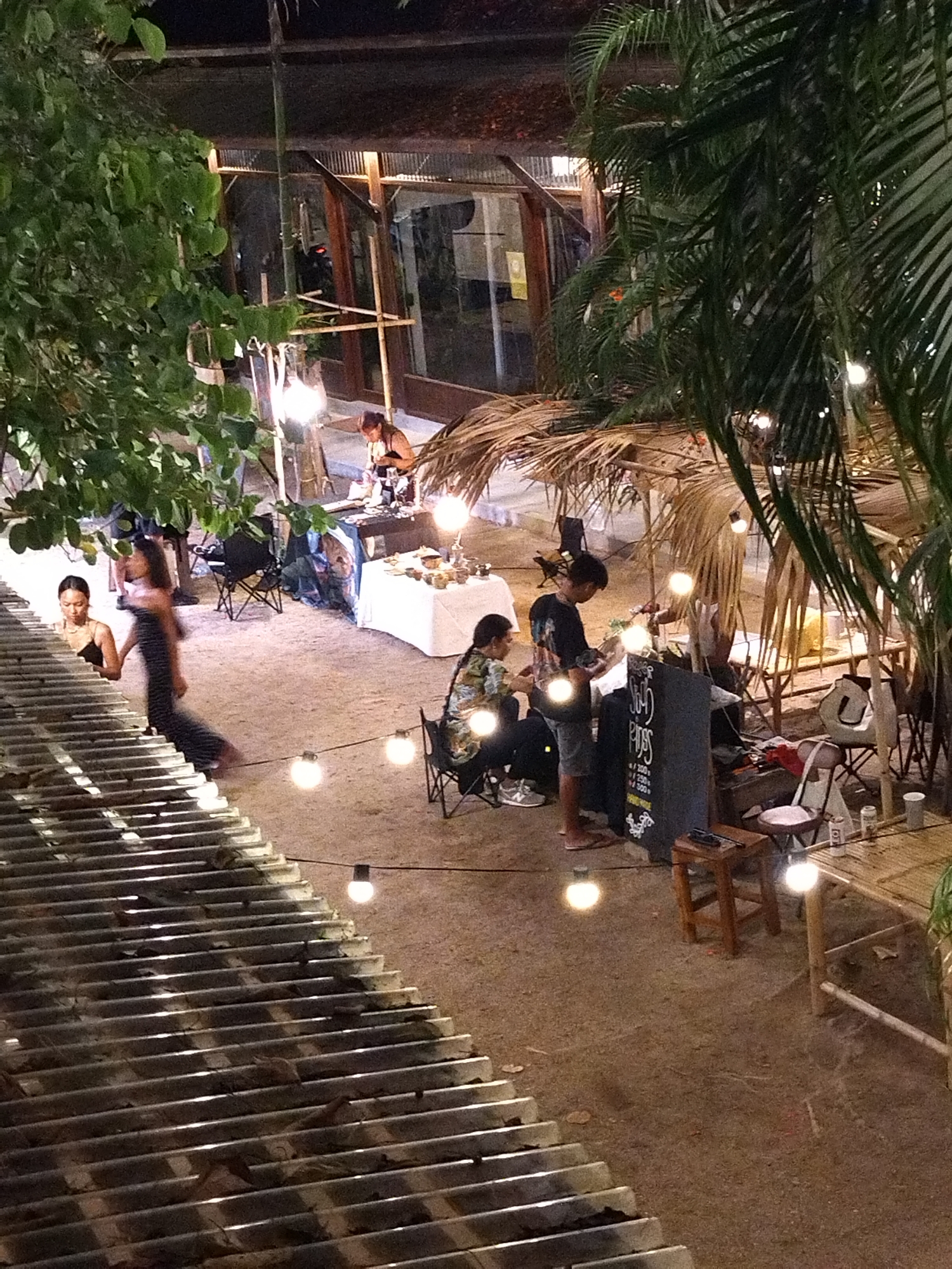 The image shows an outdoor market in ตำบลเกาะพะงัน, Thailand, at night. The market is illuminated by hanging light bulbs. People are browsing or working at the market stalls. One woman is descending a corrugated metal staircase. To the right of the staircase, a man is standing near a chalkboard sign, with a woman sitting on a chair next to him. Behind them is a stall made of straw. In the background, another woman is working at a table with goods. A tree is on the left, and palm trees are on the right.