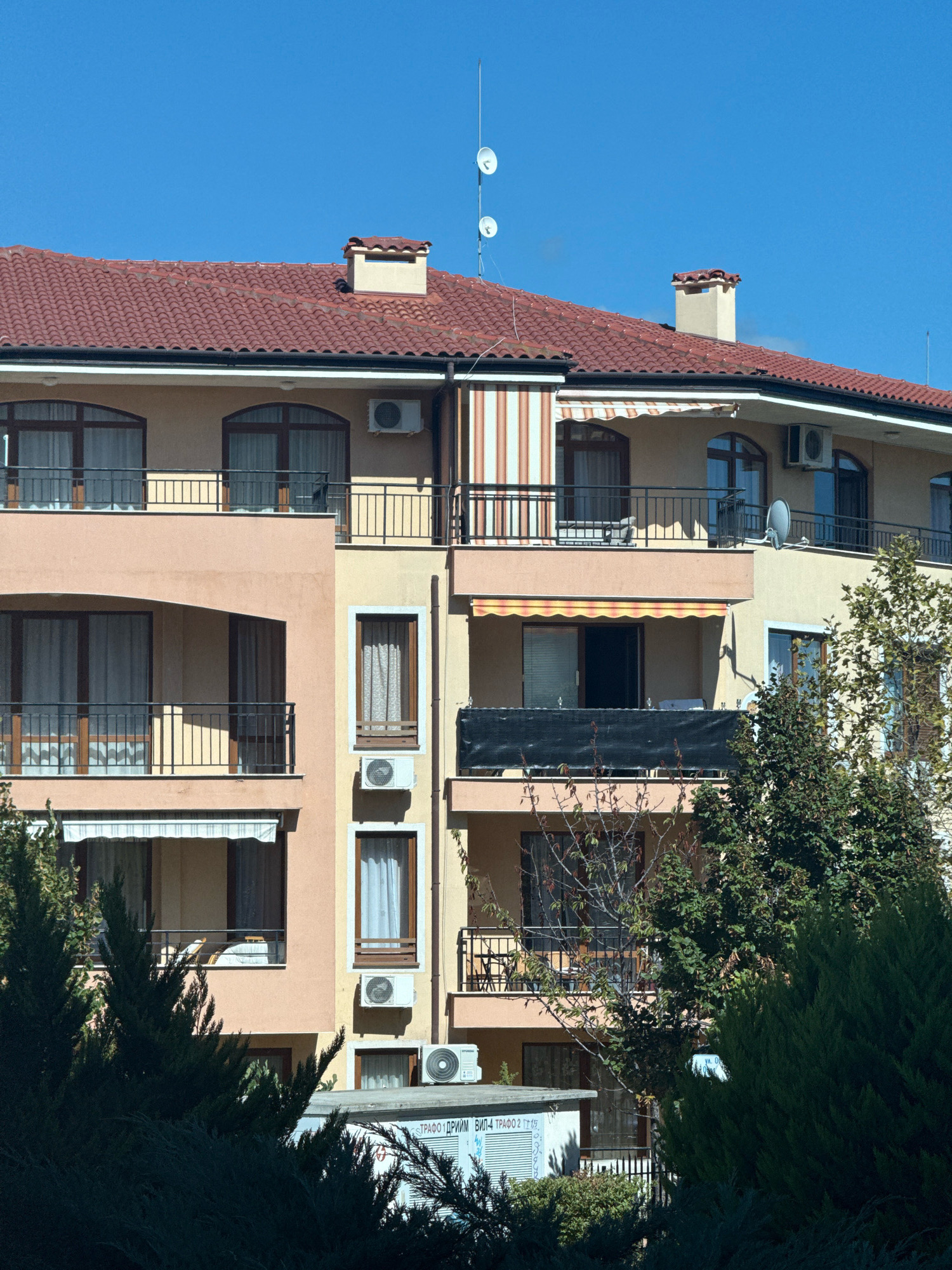 This is a daytime, eye-level exterior photograph of a multi-story apartment building in Sveti Vlas, Bulgaria. The building features a red tiled roof with two chimneys and several satellite dishes. The facade is a light peachy-orange on the left and a creamy yellow on the right. The building has multiple balconies with black railings, some with awnings. Windows are arched and rectangular, with dark brown frames and sheer white curtains visible. Several outdoor air conditioning units are mounted on the walls. Lush green trees and bushes are in the foreground and partially obscure the lower levels of the building. In the bottom of the frame, a portion of a low building or structure with a sign is visible, displaying Cyrillic text. The sky is a clear, bright blue. There are no people visible in the image.