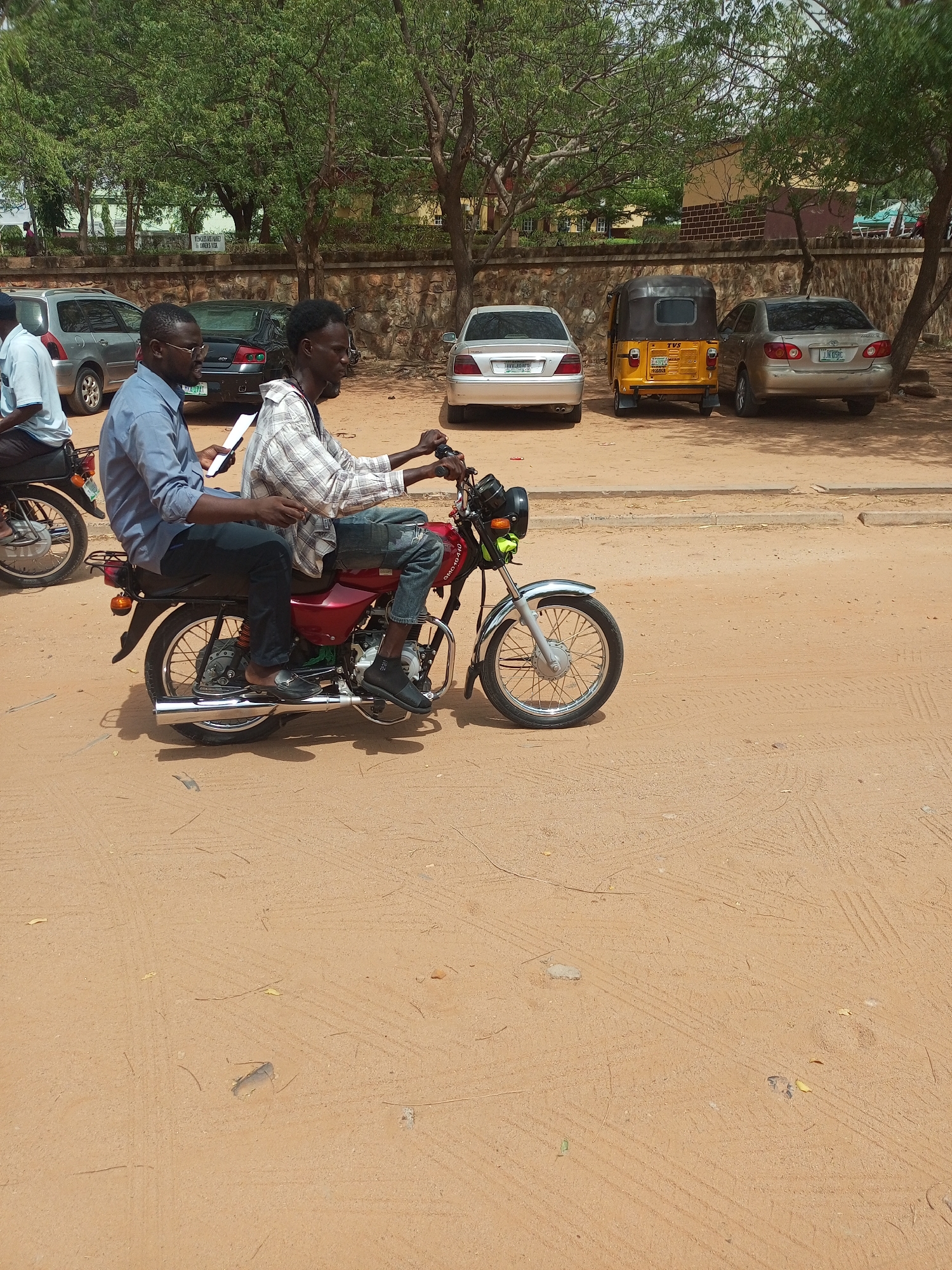 The image depicts a street scene in Gombe, Nigeria. Two men are riding a red motorcycle on a dirt road. The driver is wearing a plaid shirt and jeans, while the passenger behind him is wearing a blue shirt and black pants and holding a piece of paper. Several vehicles, including cars and a yellow tricycle, are parked along the side of the road. There is a low stone wall with trees in the background.