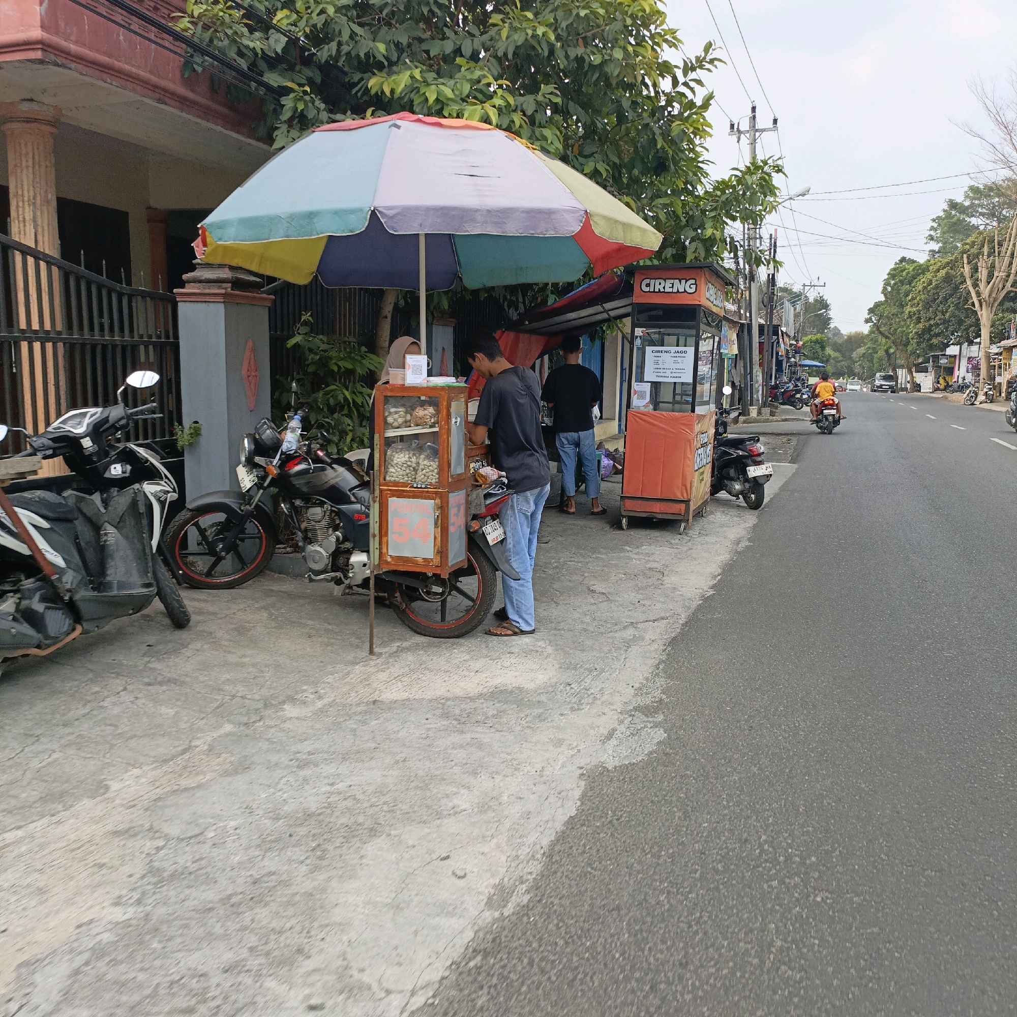 An outdoor scene shows a street in Salatiga, Indonesia, with food vendors and parked motorcycles. In the foreground, a multi-colored umbrella shades a food cart displaying snacks in a glass case. A person wearing a black t-shirt and blue jeans stands behind the cart, interacting with the vendor who is partially visible and wearing a headscarf. To the left, two motorcycles are parked on the sidewalk, one white and black, the other black with red accents on the wheels. Another food cart, orange and square, is visible further down the street. The street is paved, with a concrete sidewalk running alongside it. Trees and utility poles line the street. In the distance, several vehicles and motorcycles are seen on the road. The overall impression is of a busy street scene with local commerce.