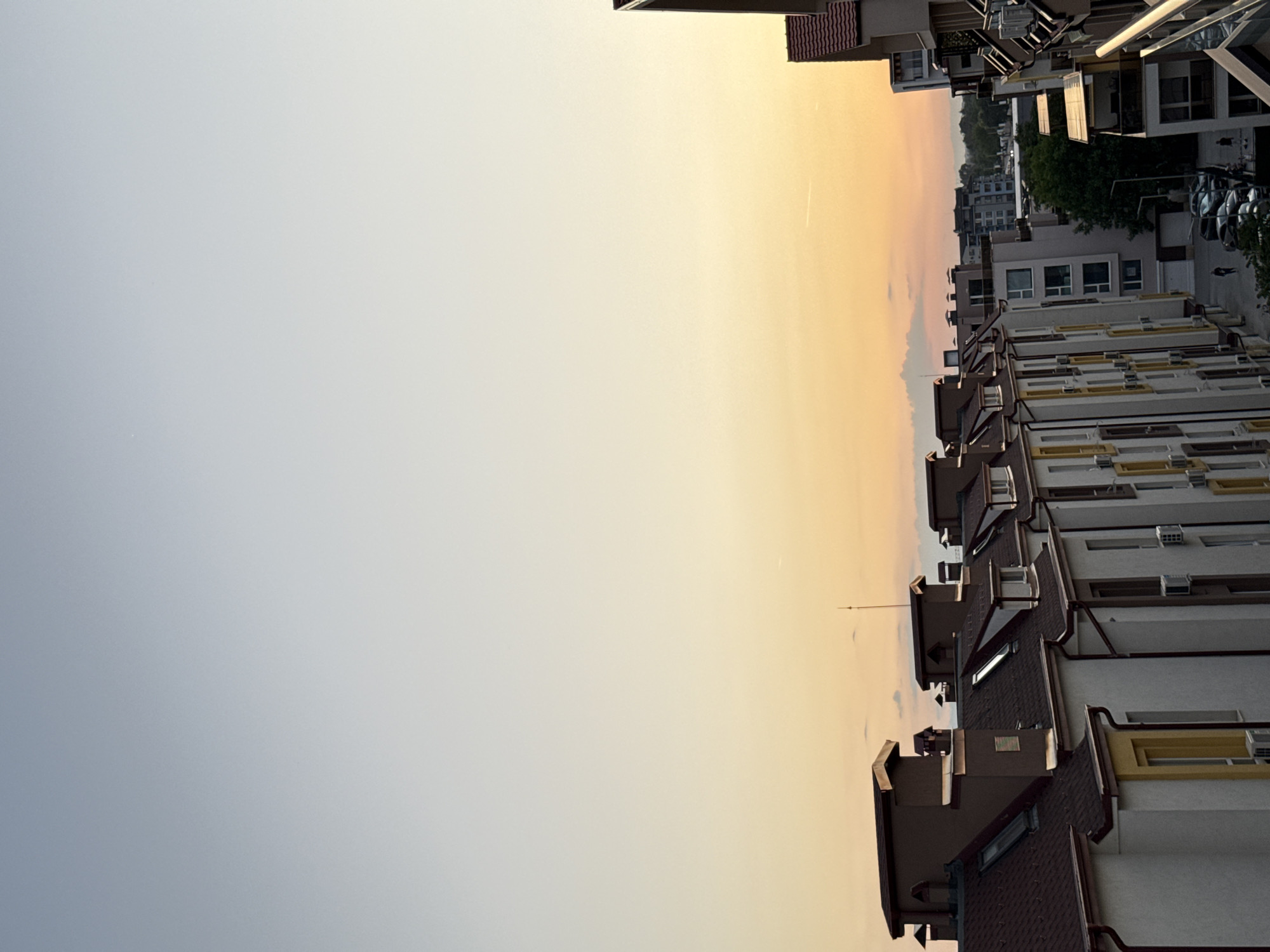 The image shows a view of a residential area in Plovdiv, Bulgaria, at dusk or dawn. The sky is a gradient of pale blue to orange, indicating the time of day. Rows of apartment buildings with brown roofs and white facades line the right side of the frame. The architecture suggests a relatively modern development. In the distance, more buildings and some greenery can be seen. The perspective implies the photo was taken from an elevated position, possibly a balcony or rooftop of another building. Cars are parked in the distance. There are no people visible.
