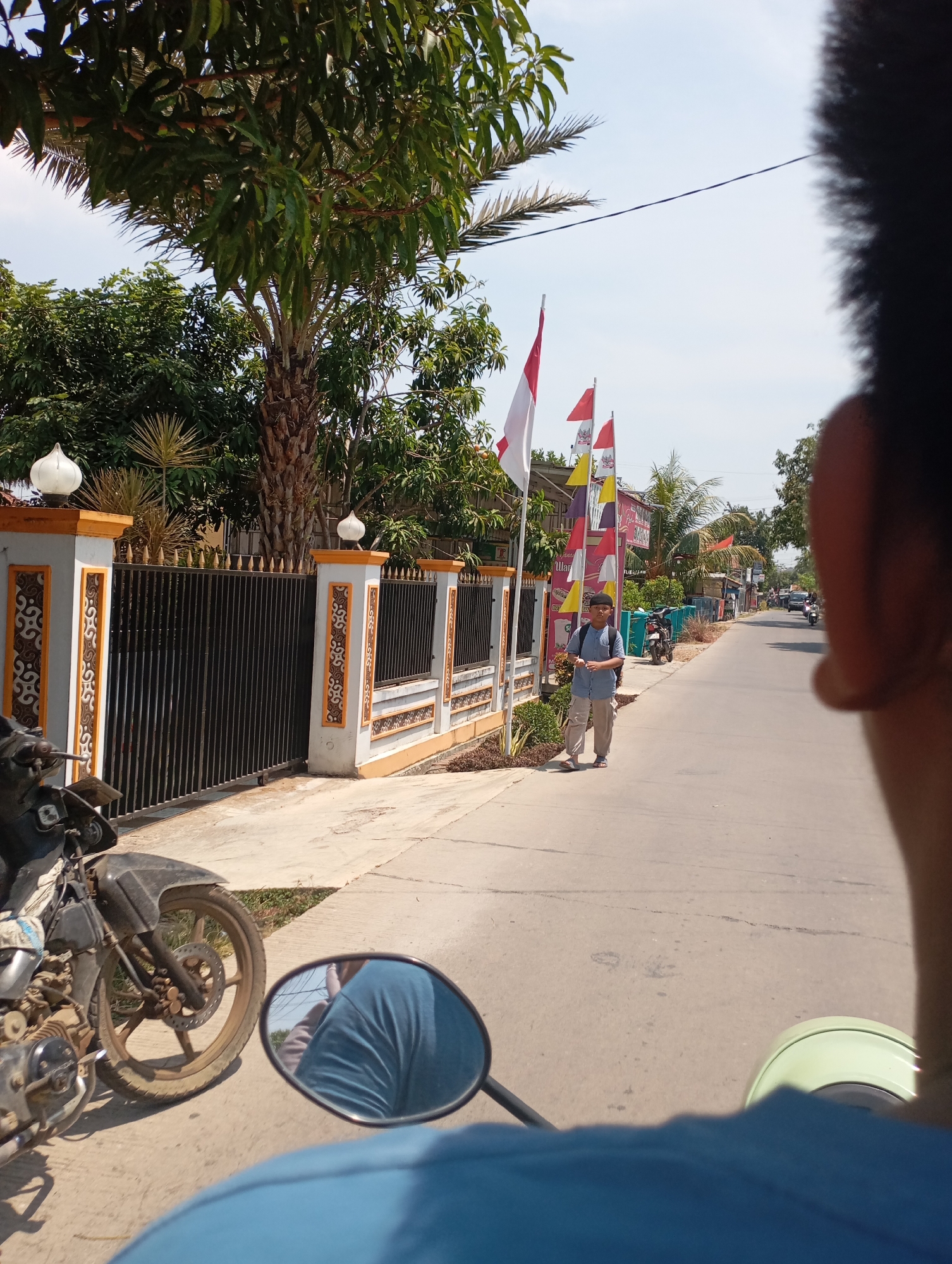 In the city of Kecamatan Haurgeulis, Indonesia, a young boy wearing a light blue shirt and light-colored pants walks on the side of a paved road. He carries a dark backpack. Several Indonesian flags, red and white, are displayed on poles behind him. The road is lined with houses and shops, and a black metal fence with ornate white and orange pillars borders the left side of the street. A parked motorcycle is visible in the foreground on the left, and another motorcycle is seen further down the road. The sky is clear and blue. The perspective suggests the image was taken from a moving motorcycle, with part of the rider's head and shoulder visible in the right foreground, and the motorcycle's handlebar and mirror in the bottom left.