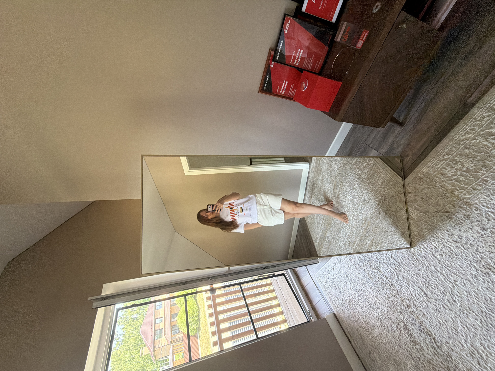 A full-body mirror reflects a young woman standing on a cream-colored rug in a room with beige walls. She is wearing a white t-shirt with a graphic design and white shorts, and is holding a smartphone in her right hand, taking a selfie. A large, open sliding glass door with a wooden railing and a view of a house and trees is to the left of the mirror. To the right of the mirror, on a dark wood dresser, are several red award plaques and a red box. The floor is a mix of the cream rug and dark wood laminate. The scene appears to be an indoor residential setting, possibly in Malinovka, Russia, given the context provided.
