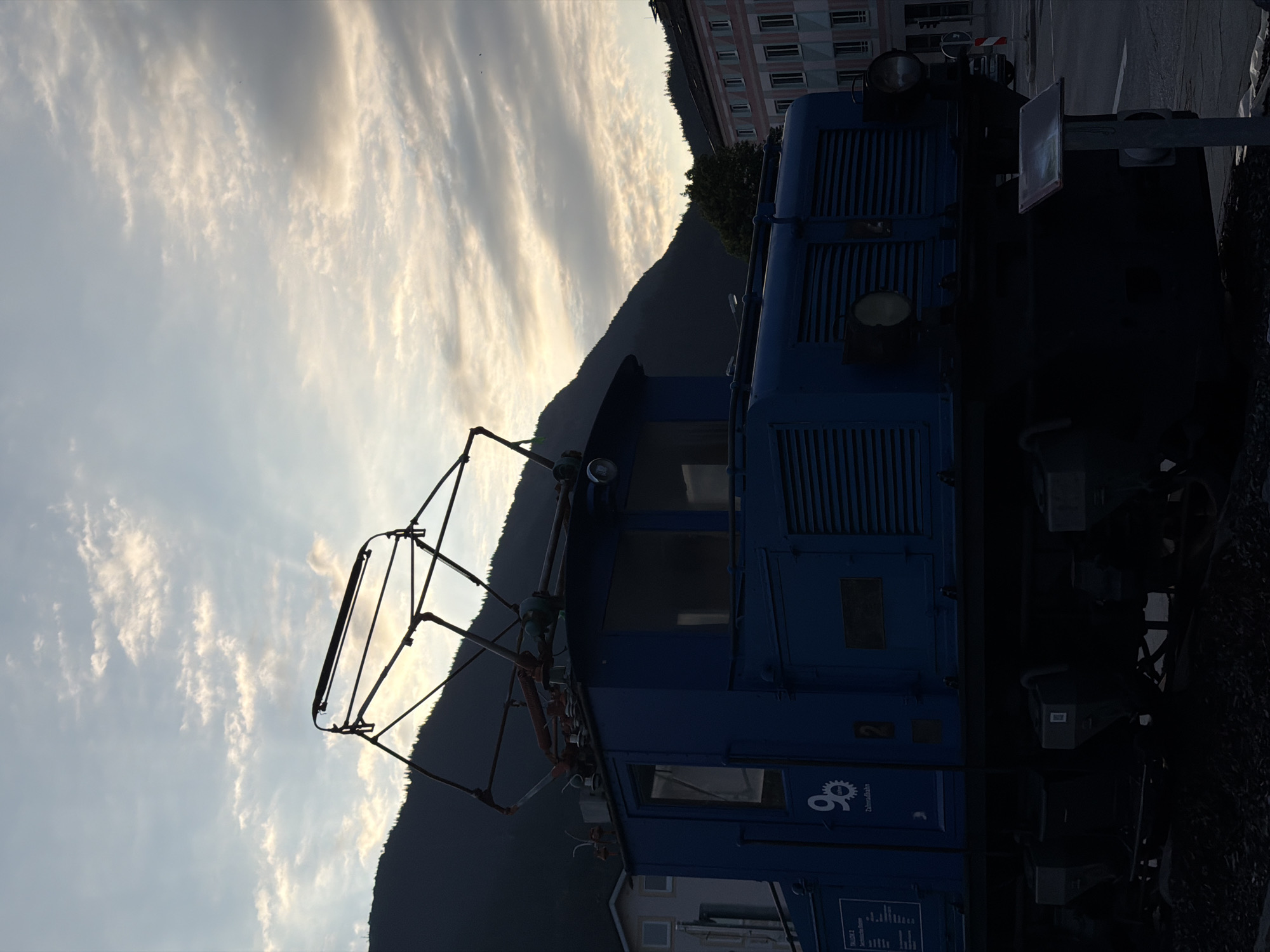An outdoor, low-angle, vertical-format shot captures the front of a dark blue electric locomotive silhouetted against a cloudy sky and mountainous background. The locomotive, positioned on tracks, has a prominent pantograph on its roof and two headlights on its front. To the left of the locomotive, a light-colored building is visible, and to the right, a multi-story building with an ornate facade stands behind a tree. The sky is filled with elongated, textured clouds illuminated by the setting sun, creating a dramatic, atmospheric effect. The scene appears to be located in a town or city with a mountainous backdrop, possibly Garmisch-Partenkirchen, Germany, given the visual context of a historic locomotive often displayed in such Bavarian towns. No people are present in the frame.