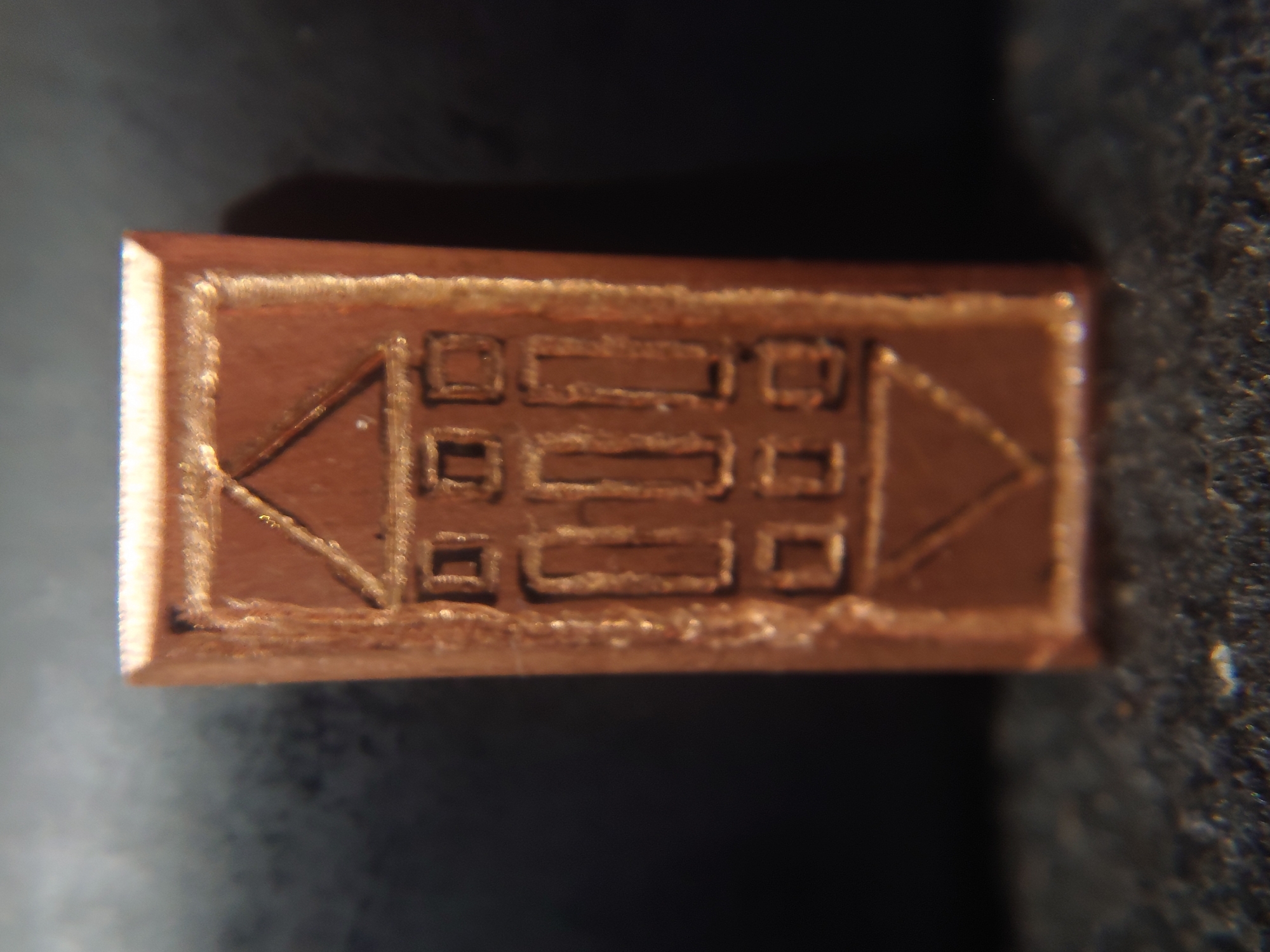 The image shows a close-up, eye-level view of a small, rectangular, copper-colored object with an engraved design. The design appears to be a stylized depiction of a building, possibly a house or a temple, with a triangular roof and multiple rectangular openings. The object is oriented vertically and has a metallic sheen. The background is dark and out of focus, with a slightly textured surface in the foreground that is also out of focus. The object is centrally framed in the image. No specific location within Sokolohirsk, Ukraine, can be determined from this image.