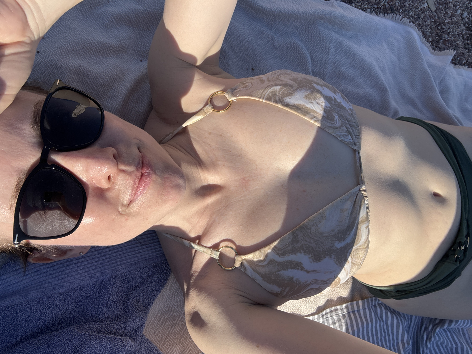 A woman is lying on a towel on a beach, wearing a bikini and sunglasses. The image is taken from a first-person perspective, looking down at the woman's torso and face. The woman is lying on her back, with her head slightly tilted to the side. She is wearing a light-colored bikini top with a marble-like pattern and a dark green bikini bottom. Her eyes are hidden behind dark sunglasses. The sun is shining brightly, casting shadows on her skin and the towel. In the background, there is a striped towel and a pebbly beach. The woman appears relaxed and is likely enjoying the sun. The location is the beach in Sveti Stefan, Montenegro.