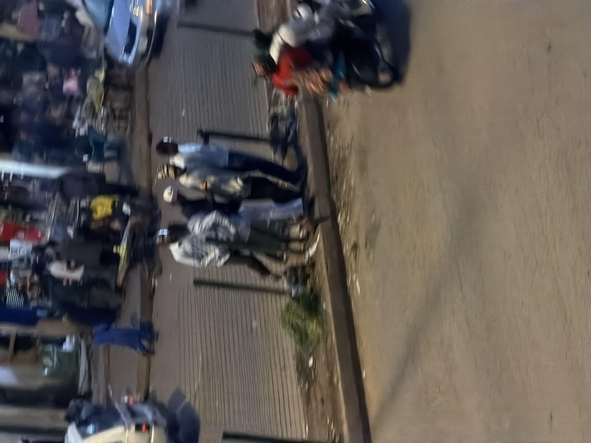 In an outdoor urban setting, likely a street in Bukuru, Nigeria, a group of individuals are visible in the mid-ground and foreground. A person in a blue garment is kneeling on the ground on the left side of the image. To their right, a group of at least four individuals stand together, appearing to be conversing or observing something. They are dressed in casual attire. A motorcycle with at least two individuals on it is in motion in the right foreground, moving away from the viewer. The background depicts a bustling street scene with buildings, stalls, and other vehicles, suggesting a market or commercial area. The ground appears to be paved, with some debris and vegetation visible. The lighting suggests it is either dawn, dusk, or nighttime.