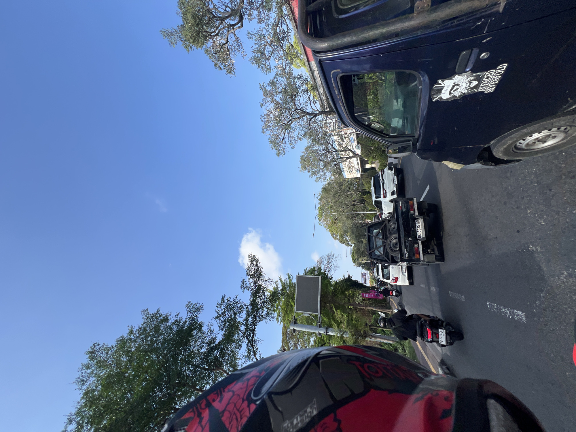 This is a first-person perspective image taken from a motorcycle in traffic on a sunny day in Denpasar, Indonesia. The rider is wearing a red and black helmet, partially visible in the foreground. The scene depicts a busy street with several vehicles including a dark blue pickup truck with an "Avenged Sevenfold" sticker on the door in the right foreground, a black pickup truck carrying spare tires, a white SUV, and several motorcycles. A man in a black hoodie is riding a scooter in front of the motorcycle the picture is taken from. Tall trees line the side of the road, and there is a traffic sign visible on a pole. The sky is clear blue with a few wispy clouds. The road surface is asphalt with white lane markings. Buildings and advertisements are visible in the background.