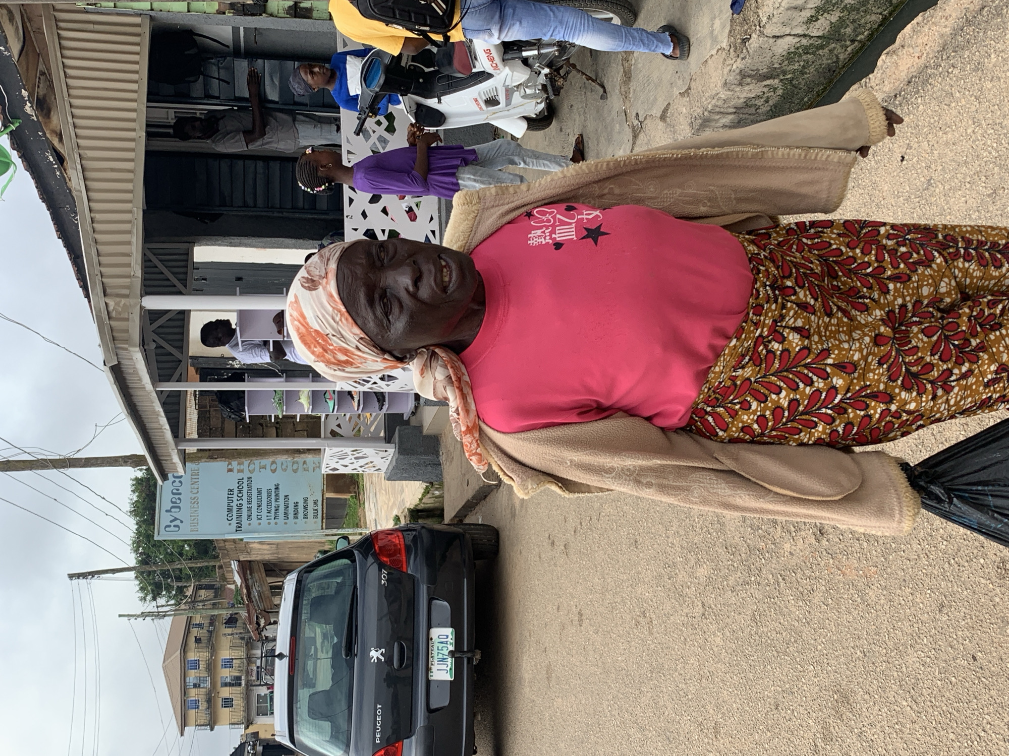 A low-angle, medium shot captures a street scene in Jos, Nigeria. An elderly woman with dark skin, wearing a pink t-shirt, a patterned skirt, and a headscarf, walks towards the right. She holds a black plastic bag in her left hand. Behind her, a black Peugeot 307 car is parked on the left side of the road. A building with a corrugated metal roof and an open front stands on the right, with a white motorcycle parked outside. A sign for "Cybercorp Business Centre" is visible on the left side of the building. Two young girls and two men are also present, with one girl in a purple top and the other in a blue t-shirt. The ground is paved, and the sky is overcast.