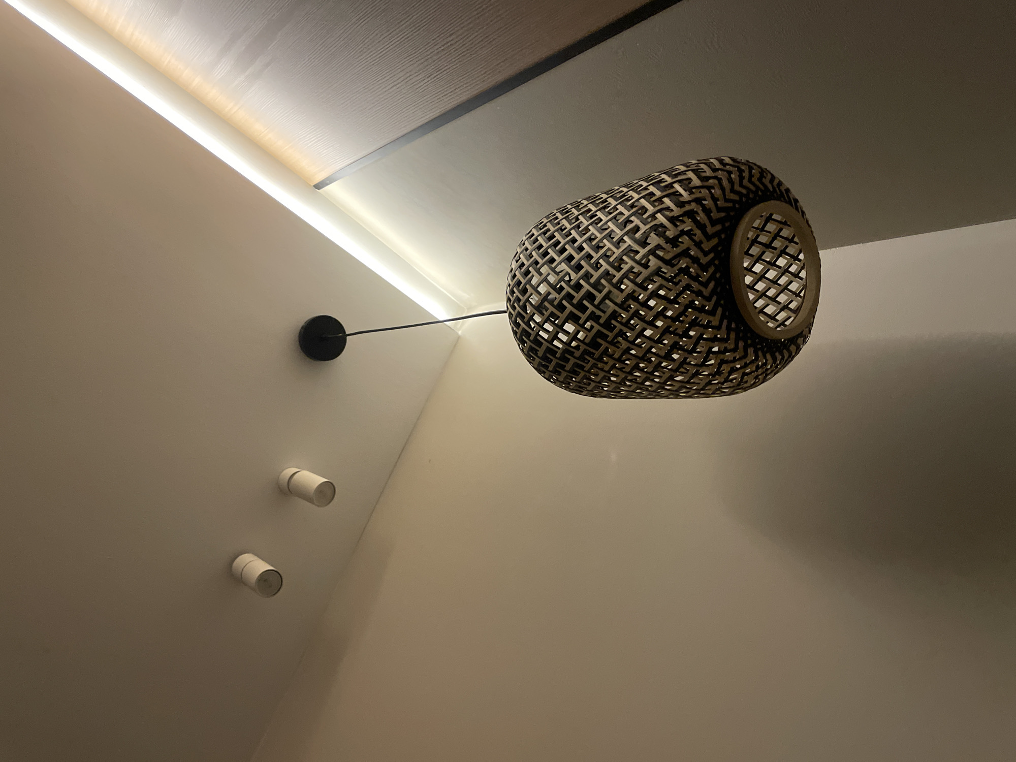 The image depicts an interior scene. A woven, cage-like lamp hangs from the ceiling on a thin black wire connected to a circular mount on the wall. Two cylindrical spotlights are visible on the wall below the lamp mount. The ceiling has recessed lighting, and the walls are off-white. The location appears to be in a room with modern interior design, possibly in a residential or commercial building. The image does not contain any people or specific actions. The location in the city of Πόρτες, Greece is not explicitly shown.