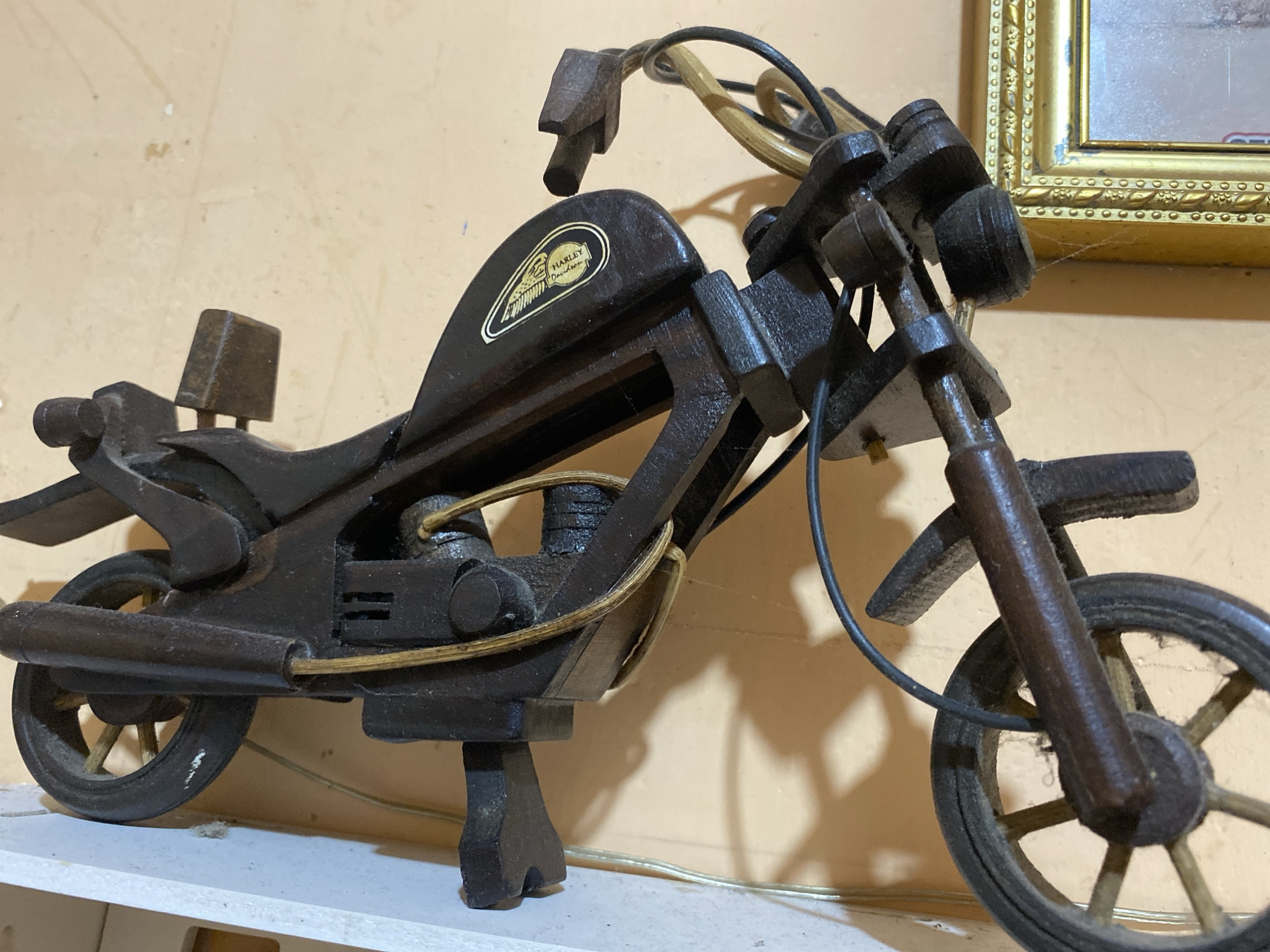 A wooden model motorcycle, possibly a Harley-Davidson due to a visible emblem, is displayed indoors. The motorcycle is dark brown and features wire details for accents. It is positioned on a white surface against a light beige wall. A gold-trimmed picture frame is visible in the upper right corner, and a thin layer of dust or cobwebs can be seen on the motorcycle and in the background. The lighting suggests an indoor setting, possibly a home or shop. The location is Unknown, Unknown.