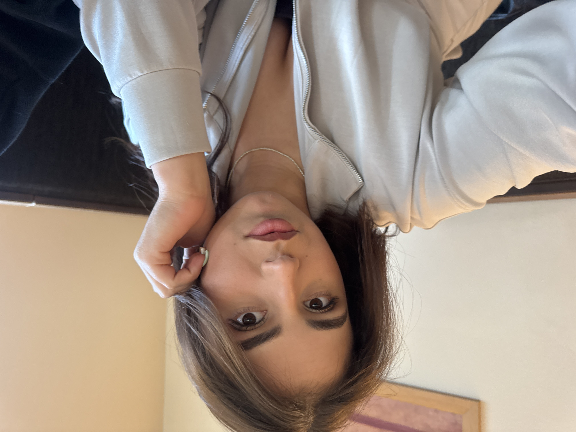 The image is a selfie taken from a low angle looking up at a woman with light brown hair, brown eyes, and fair skin. She is wearing a beige zip-up hoodie. A thin silver necklace is visible around her neck. Her left hand is near her face. The background appears to be an indoor setting with beige walls. Without additional information, determining the specific location in Sofia, Bulgaria, is impossible.