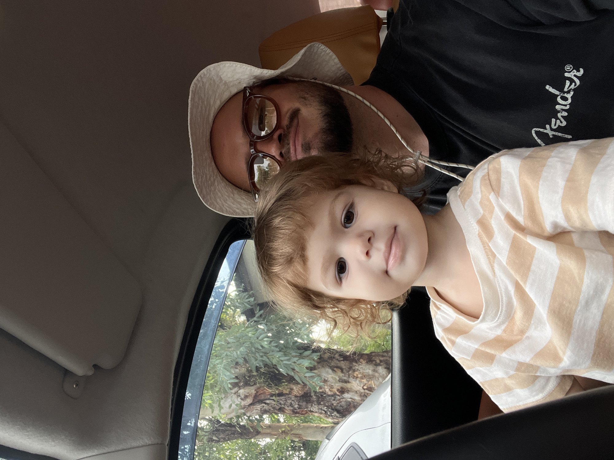 A medium shot shows a father and child in the passenger seats of a car. The child, in the foreground and slightly to the left, has light brown curly hair and is wearing a striped t-shirt. The father, to the right, is wearing a white bucket hat, sunglasses, and a black t-shirt with a "Fender" logo. They appear to be on a road trip, with trees and foliage visible through the car window. The interior of the car, including the sun visor and car seat, is also visible. The lighting suggests it is daytime.