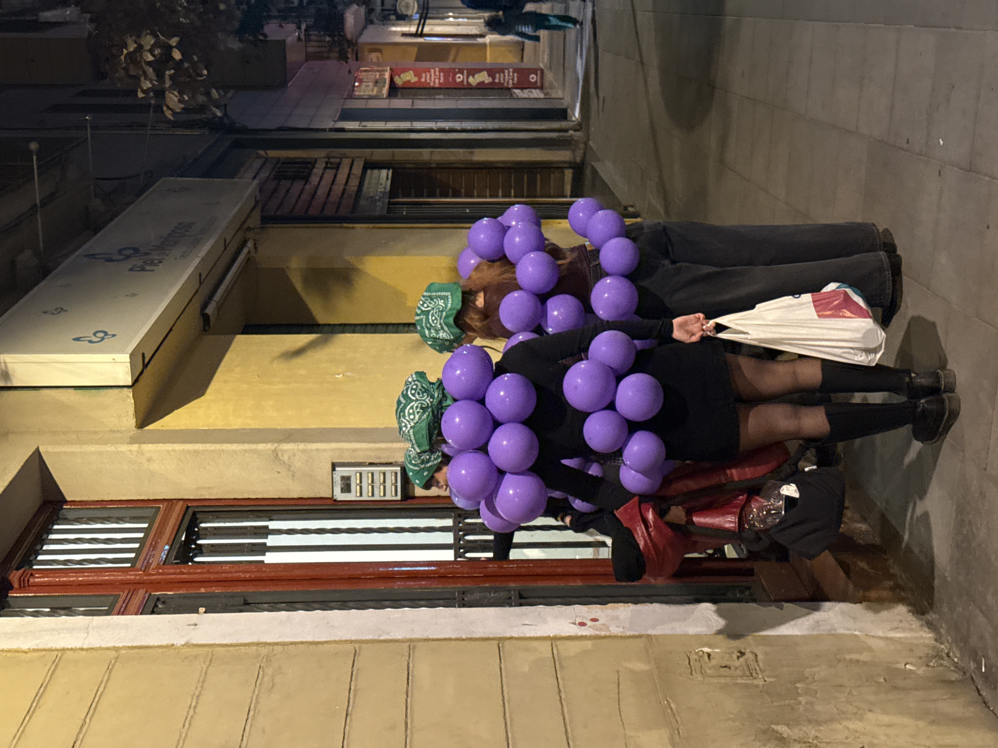 Two people are dressed as grapes, covered in purple balloons. They are wearing green bandanas and standing on a sidewalk in Barcelona, Spain, as indicated by the building architecture. One person wears a black dress and the other dark pants. One holds a white plastic bag. The scene is at night on a street with buildings and some pedestrian traffic in the background.