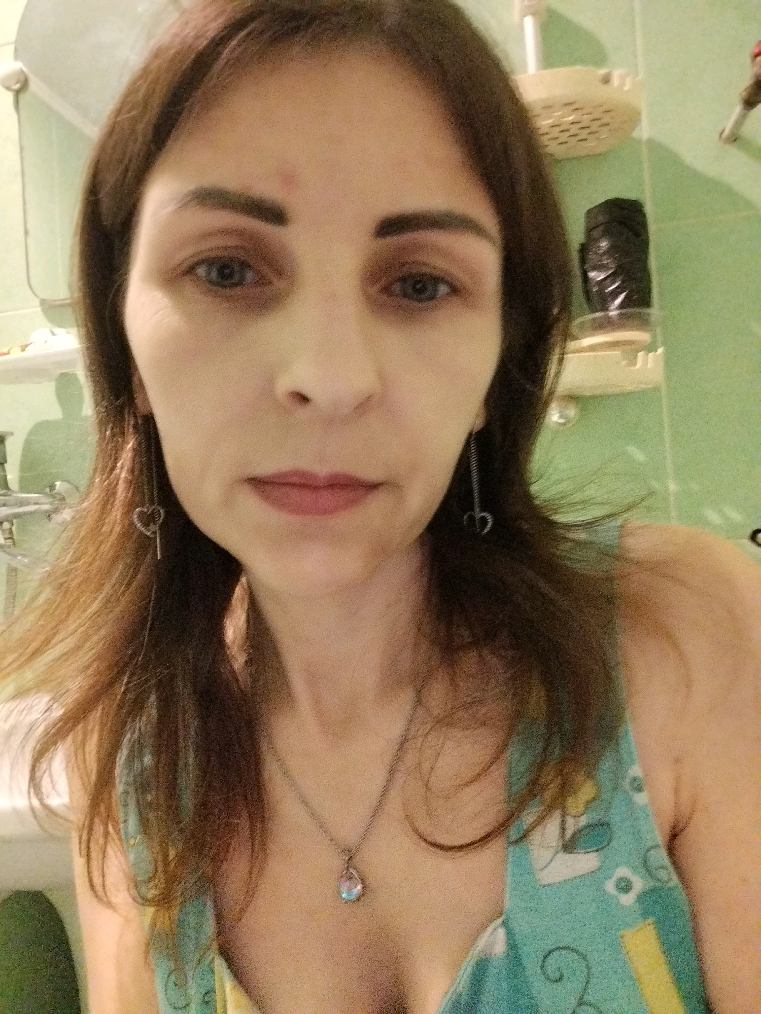 This is a close-up selfie of a woman with medium-length brown hair, taken indoors. She is wearing a light-colored necklace with a pendant and dangling earrings shaped like keys with hearts. Her shirt has a floral pattern. The background appears to be a bathroom, with a tiled wall and shelves visible. The lighting suggests it may be artificial. There is no specific indication of the location being in Vinnytsia, Ukraine.