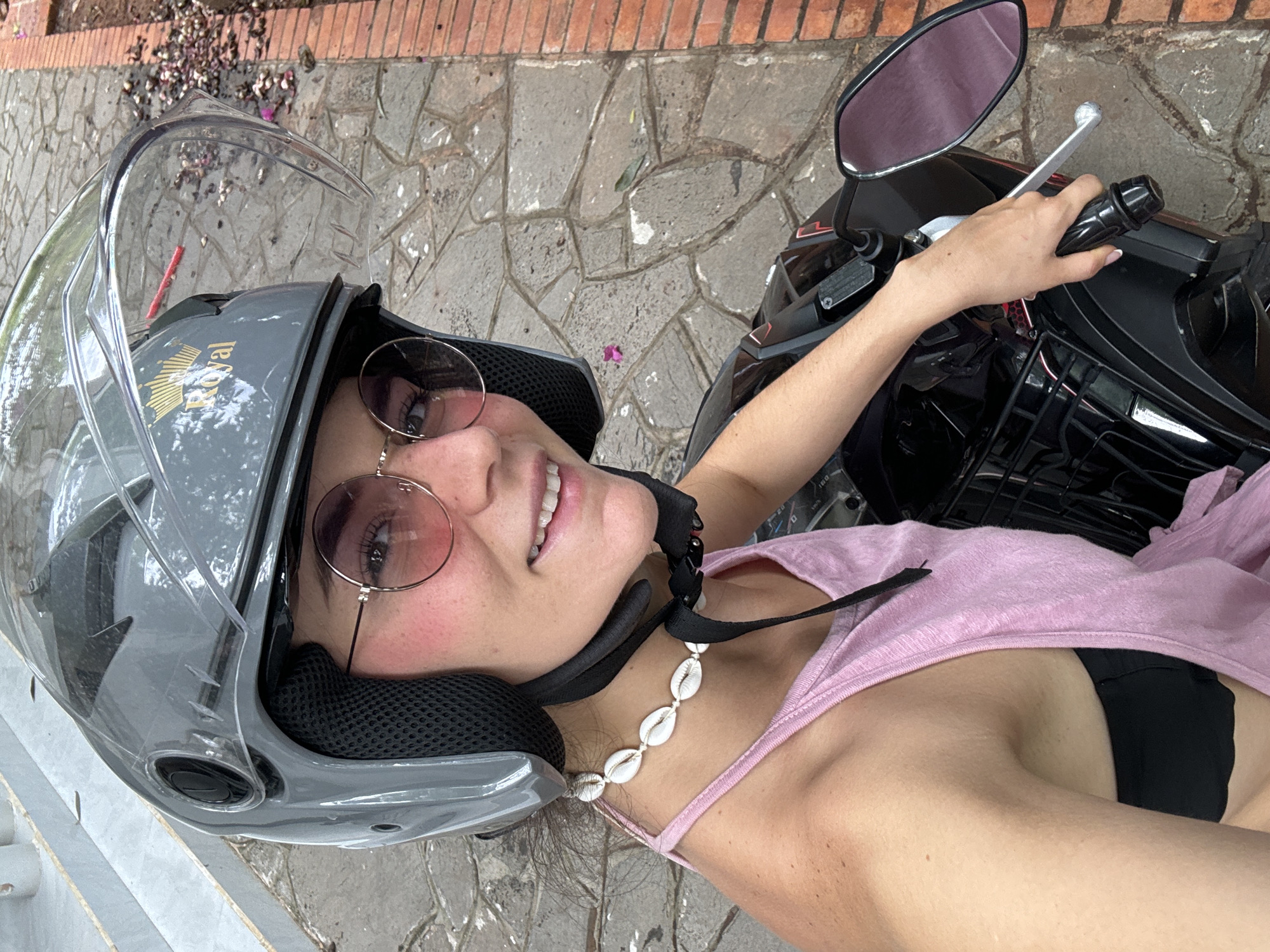 A high-angle, portrait-oriented shot captures a young woman from the chest up, smiling cheerfully while appearing to be on a black scooter. She wears a gray open-face helmet with a clear visor, which features a gold "Royal" logo on its side. Her eyes are covered by round, pink-tinted sunglasses. A distinctive necklace made of white cowrie shells hangs around her neck, and she is dressed in a light pink tank top, with a black bra visible underneath. Her left arm extends to the right, gripping the handlebar of the scooter, which also shows its rearview mirror and brake lever.
The background is a paved ground surface composed of irregular gray and brown stones, littered with scattered leaves, petals, and debris. A reddish-brown brick border lines a portion of the pathway. The natural lighting indicates it is daytime. The woman's broad smile conveys happiness and ease, suggesting a pleasant moment captured during an outing in Mũi Né, Vietnam.
