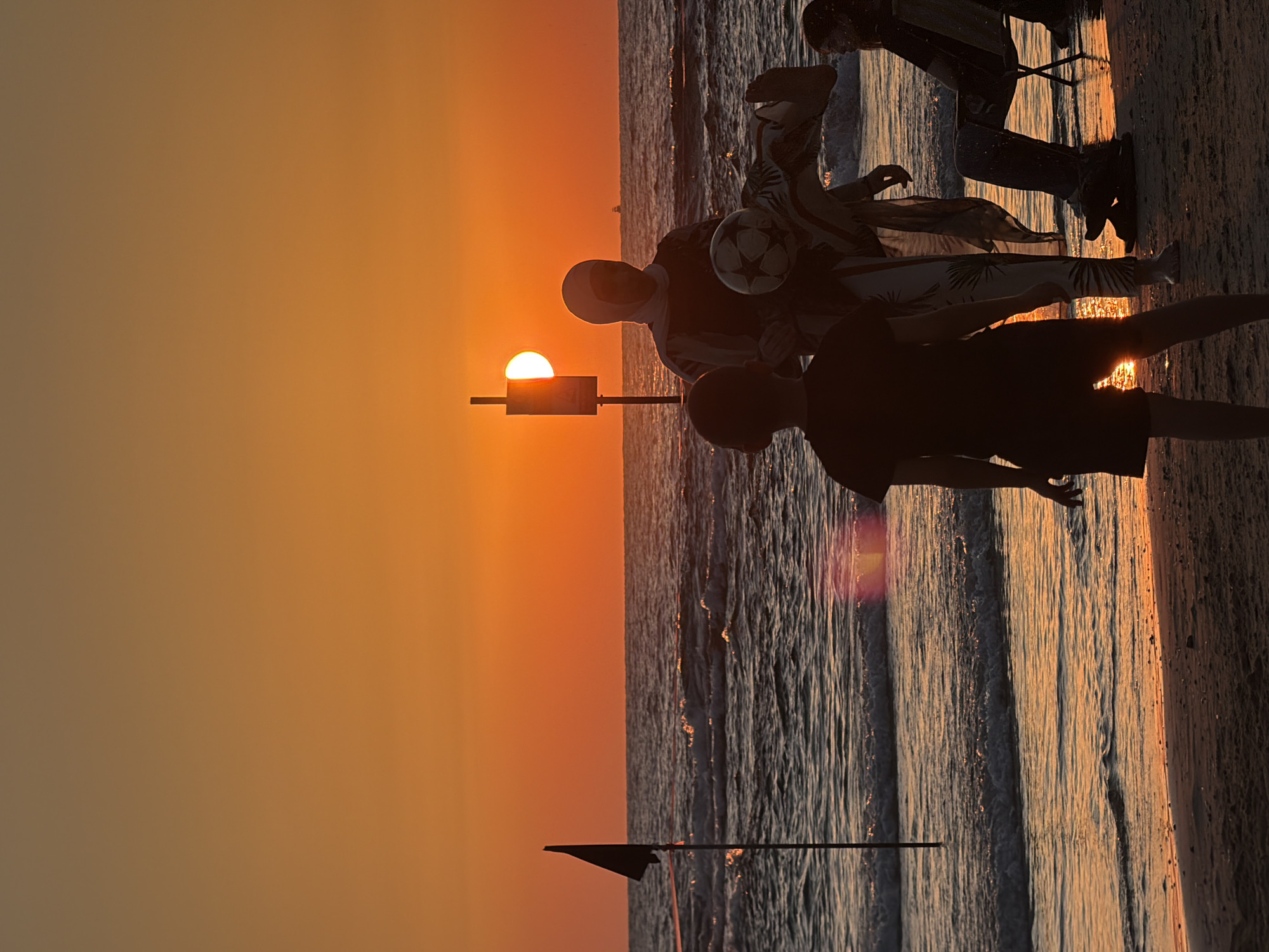 The image depicts a sunset on a beach in Tel Aviv, Israel. Silhouettes of people are visible on the sand. In the foreground, a person wearing shorts and a t-shirt is facing away from the camera. To their right, another person, possibly a woman wearing a headscarf, is standing and appears to be juggling a soccer ball with their foot. Another individual is seated in a chair to the far right. The ocean stretches out to the horizon, with waves gently lapping the shore. The sky is a vibrant orange, indicative of a sunset. A black flag on a pole stands in the water to the left of the frame.