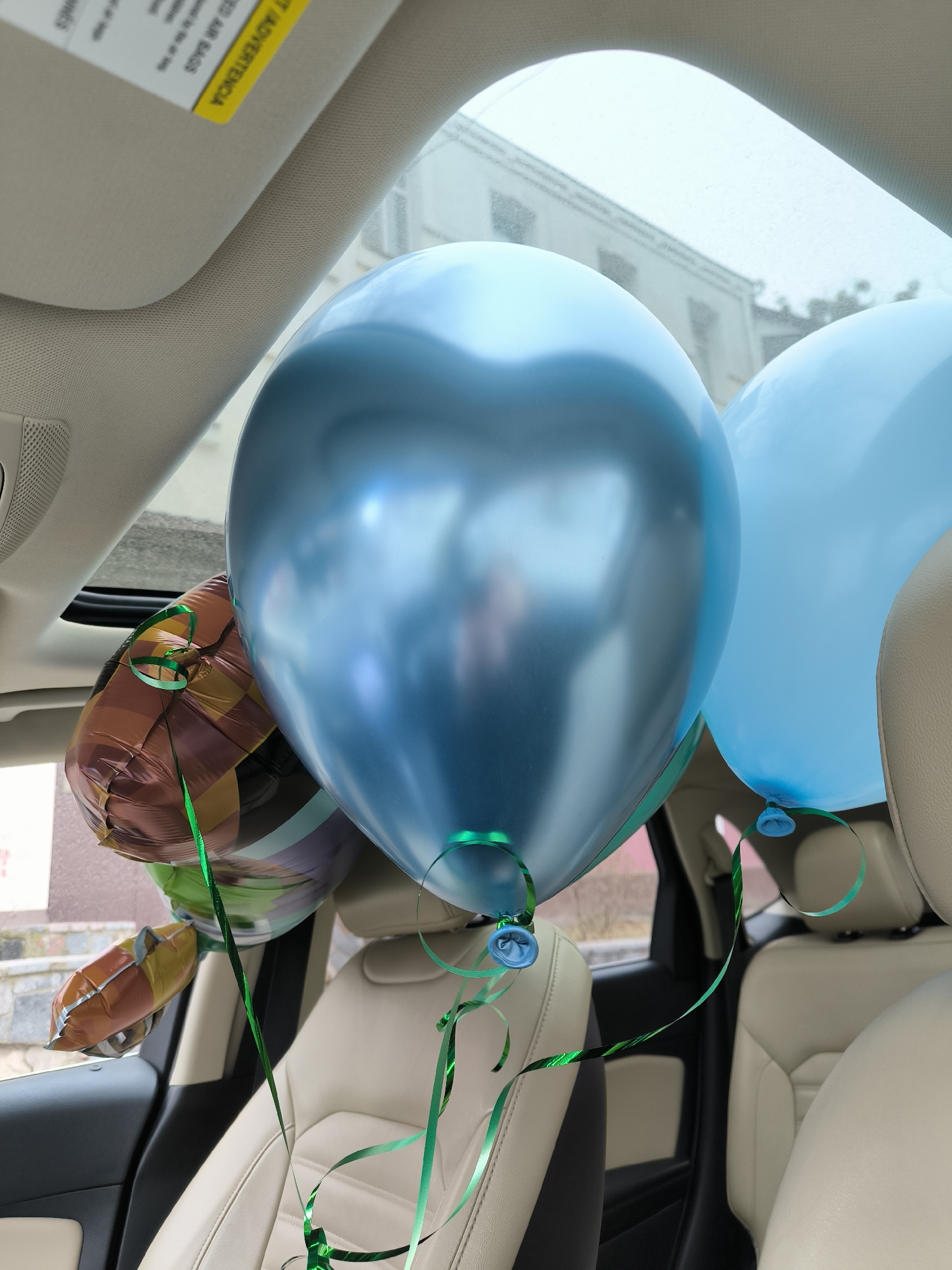 The interior of a vehicle is filled with inflated balloons. There are two blue round balloons and one foil balloon with brown, purple, and yellow patterns. Green ribbons are attached to each balloon. The car seats are beige leather. Outside the car window, a building can be seen, suggesting an urban environment. The image does not provide enough information to determine the specific location in Vinnytsia, Ukraine.