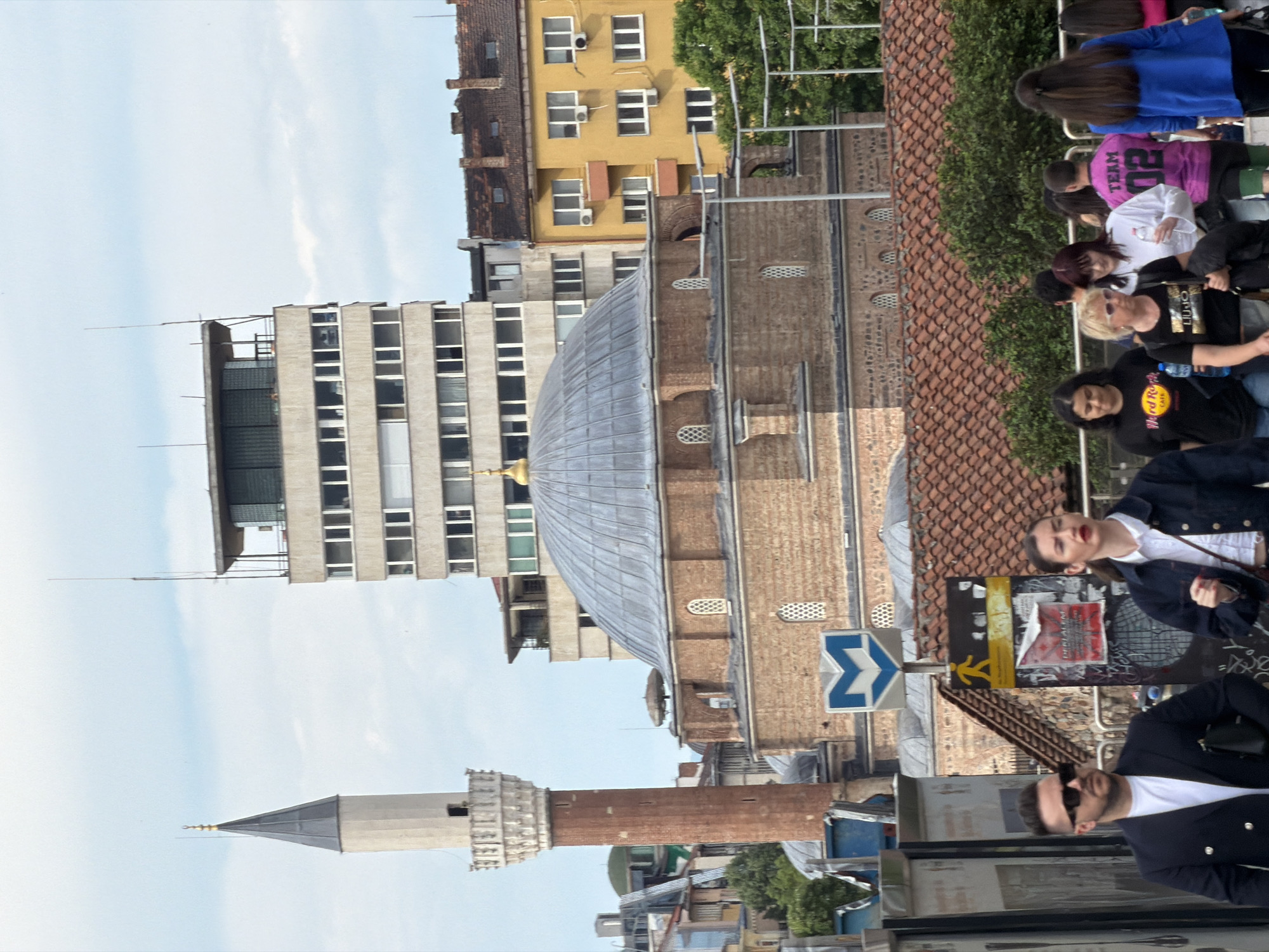 The image shows a street scene in Sofia, Bulgaria, featuring the Banya Bashi Mosque and surrounding buildings. The mosque's distinctive dome and minaret are visible, contrasting with the taller, modern building behind it. A group of people, possibly tourists, are present on the sidewalk. A road sign with a blue and white symbol can be seen, along with urban signage and street-level objects. The sky is light blue with a few clouds. The scene suggests a typical day in an urban environment with historic architecture.