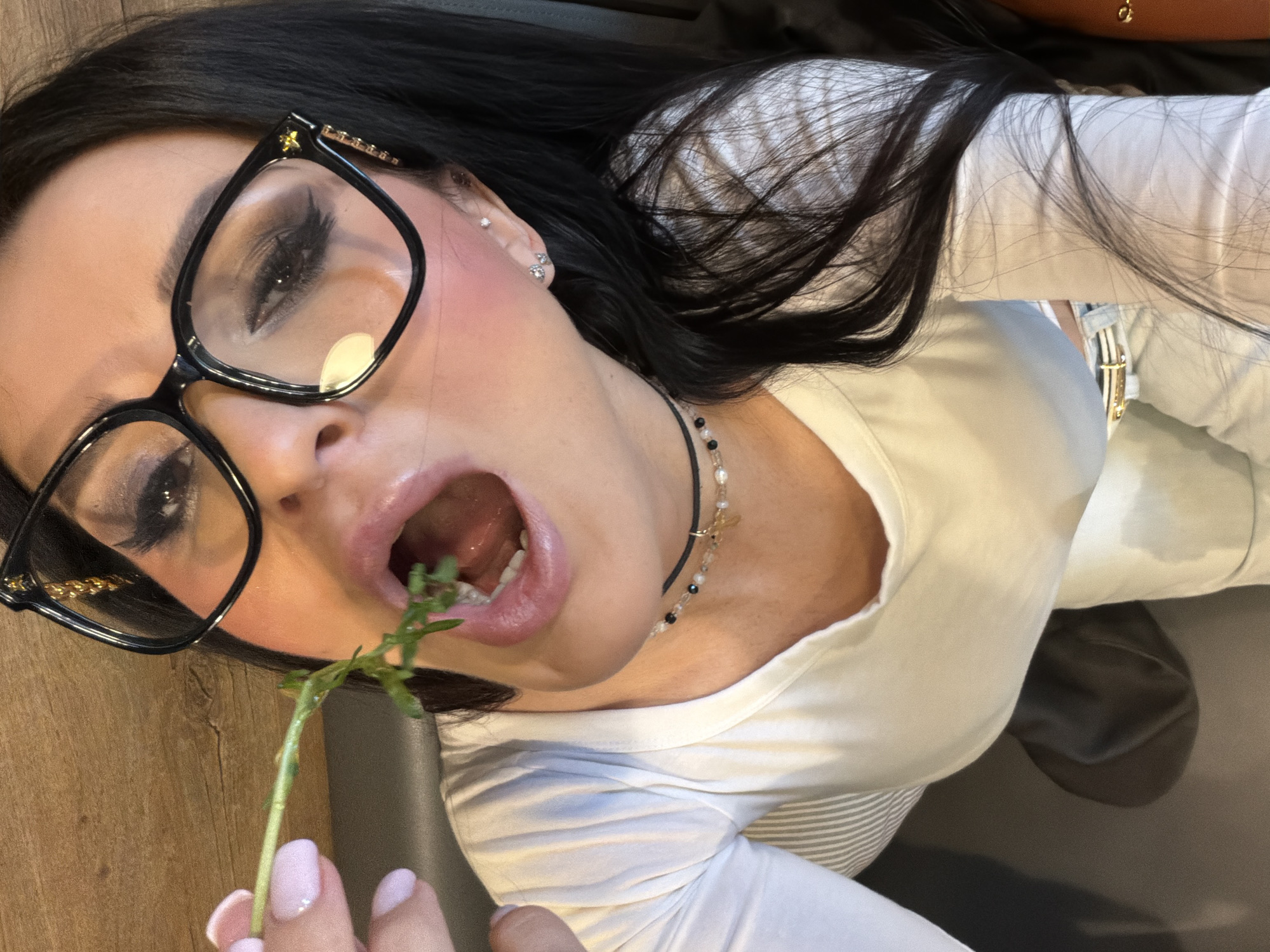 The image presents an angled, close-up portrait of a woman, focused on her head and upper torso. She has long, dark hair that cascades around her. Her face is adorned with prominent black-framed glasses, featuring a small gold star detail on the frame near her right temple. Her eyes are enhanced with dark eyeshadow and eyeliner, and a hint of blush colors her cheek. She wears multiple small, silver-toned earrings on her ear.
Her mouth is wide open, revealing her teeth and tongue, as she holds a slender green plant stem. Her fingers, with light pink polished nails, grip the stem as it enters her mouth. She is dressed in a white long-sleeved top, possibly layered over a light-colored striped garment, and a delicate black choker necklace with small beads and a charm around her neck.
The background includes a warm-toned wooden surface, likely a table or floor, and a dark, possibly upholstered surface like a chair or couch. The lighting suggests an indoor setting. The overall expression appears to be a deliberate, posed shot, possibly conveying a playful or artistic mood. Based on the provided context, the scene is located in Moscow, Russia. There is no visible text in the image.