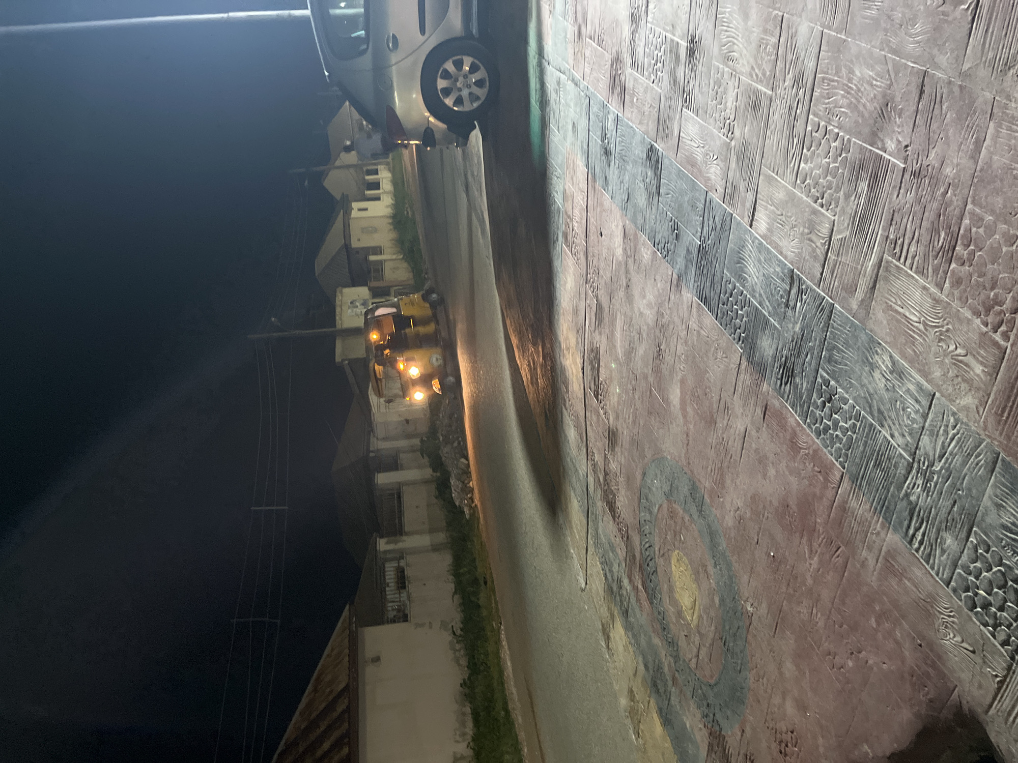 At night in Abuja, Nigeria, a yellow auto-rickshaw with its headlights illuminated travels down a paved road. To the right of the auto-rickshaw, a silver sedan is parked. The sidewalk in the foreground is paved with decorative patterned tiles. In the background, several single-story buildings with corrugated metal roofs are visible, along with a fence. The sky is dark, indicating nighttime.