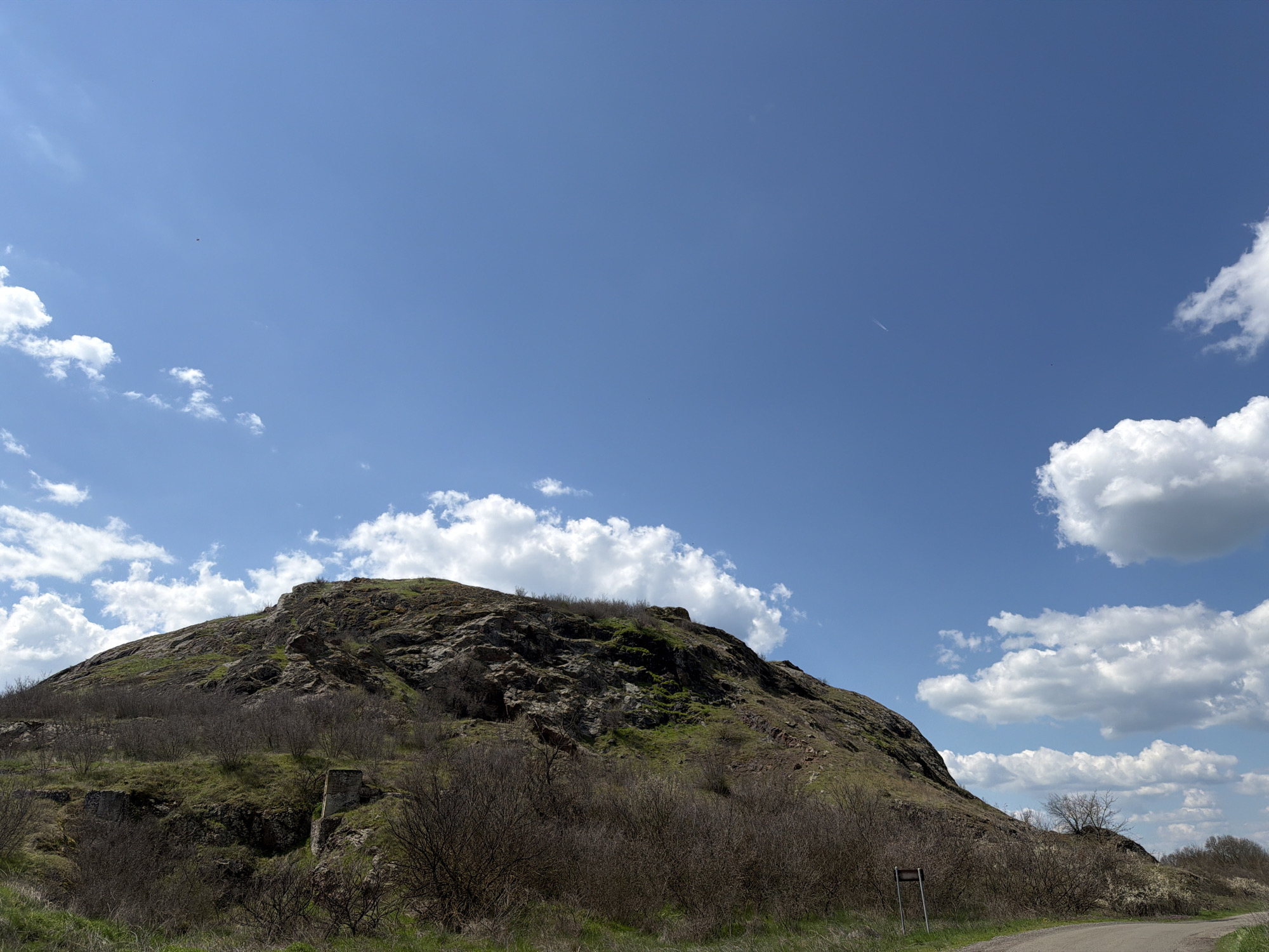 The image shows a hill under a bright blue sky with scattered white clouds. The hill is rocky, with patches of green vegetation and areas covered in dry, brown bushes. A small, seemingly man-made structure can be seen on the side of the hill, appearing as a remnant of an old construction. A road is visible at the bottom of the frame, running along the base of the hill. A signpost stands beside the road. The location is identified as Rusokastro, Bulgaria, based on prior knowledge.