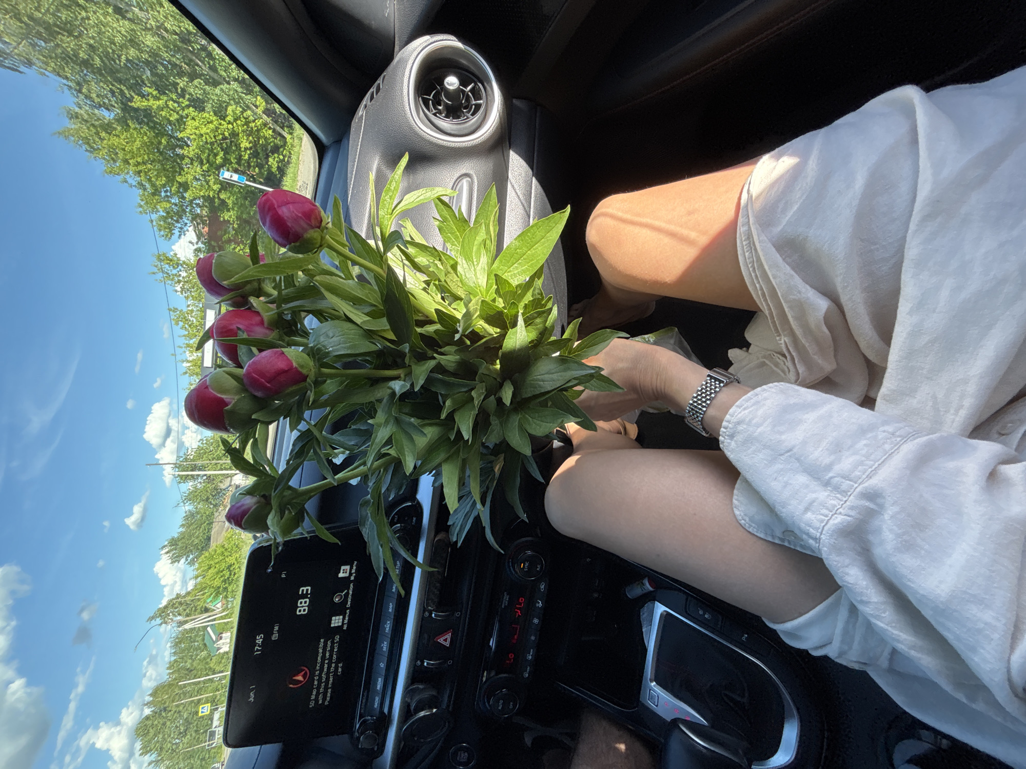 Here is a description of the image:
The interior of a car is shown, with a person wearing a white shirt and a metal watch visible in the passenger seat. The dashboard is black and features a digital display showing "Jun, 1", "17:45", "FM1", and "88.3" alongside a warning message regarding an incompatible SD map card. A bouquet of red peonies is placed between the dashboard and the passenger, with greenery filling much of the space. Outside the car window, trees and a blue sky with some clouds are visible. Based on the text on the display, this image was taken on June 1, 2023. The location is not specified in the picture.