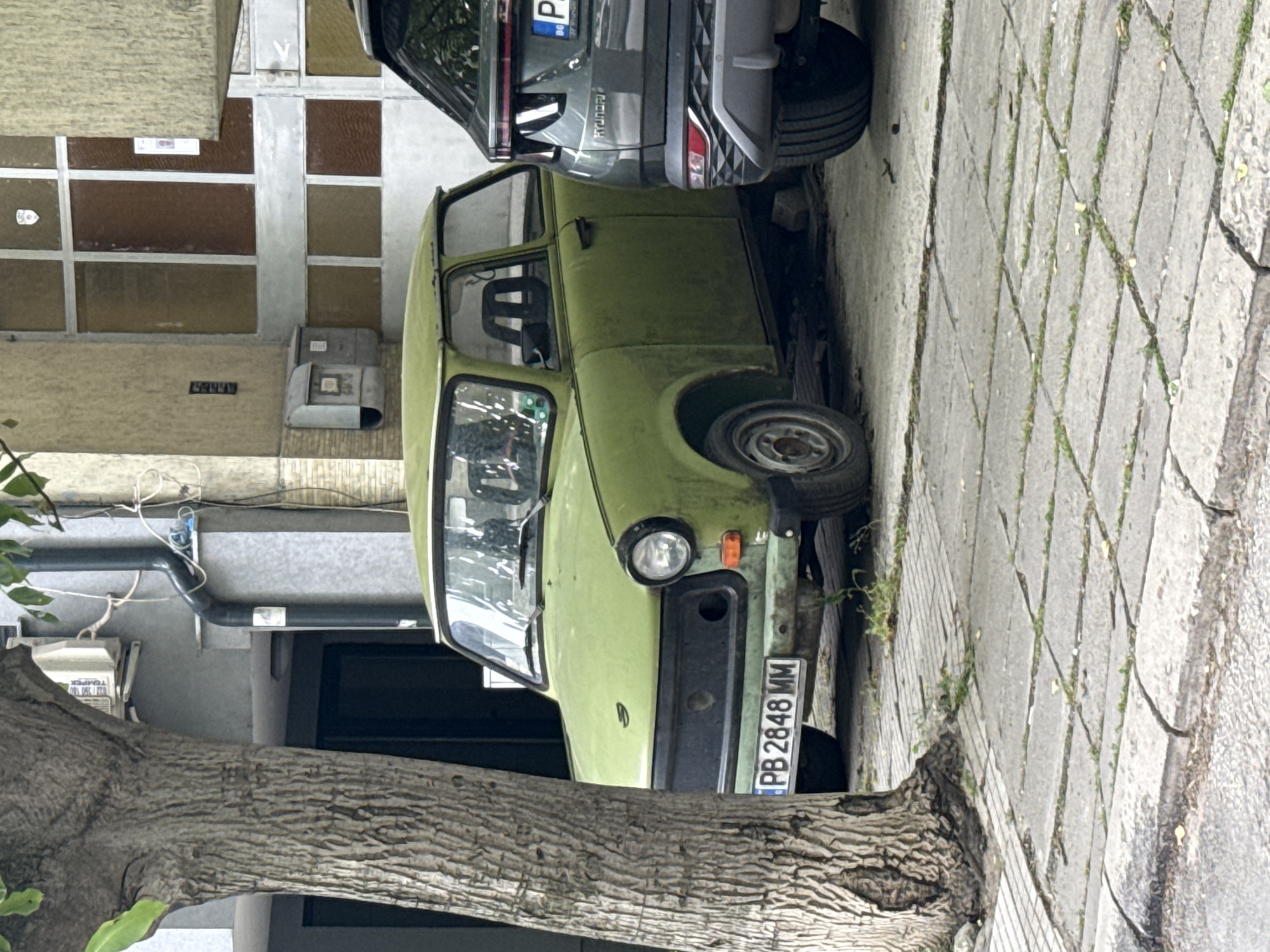 An aged, light green Trabant automobile is parked adjacent to a modern, dark-colored Hyundai SUV on a paved sidewalk in Plovdiv, Bulgaria. The Trabant has a license plate that reads "PB 2848 MM". The cars are positioned in front of a building with a partially visible exterior. A tree trunk is situated near the front of the Trabant.