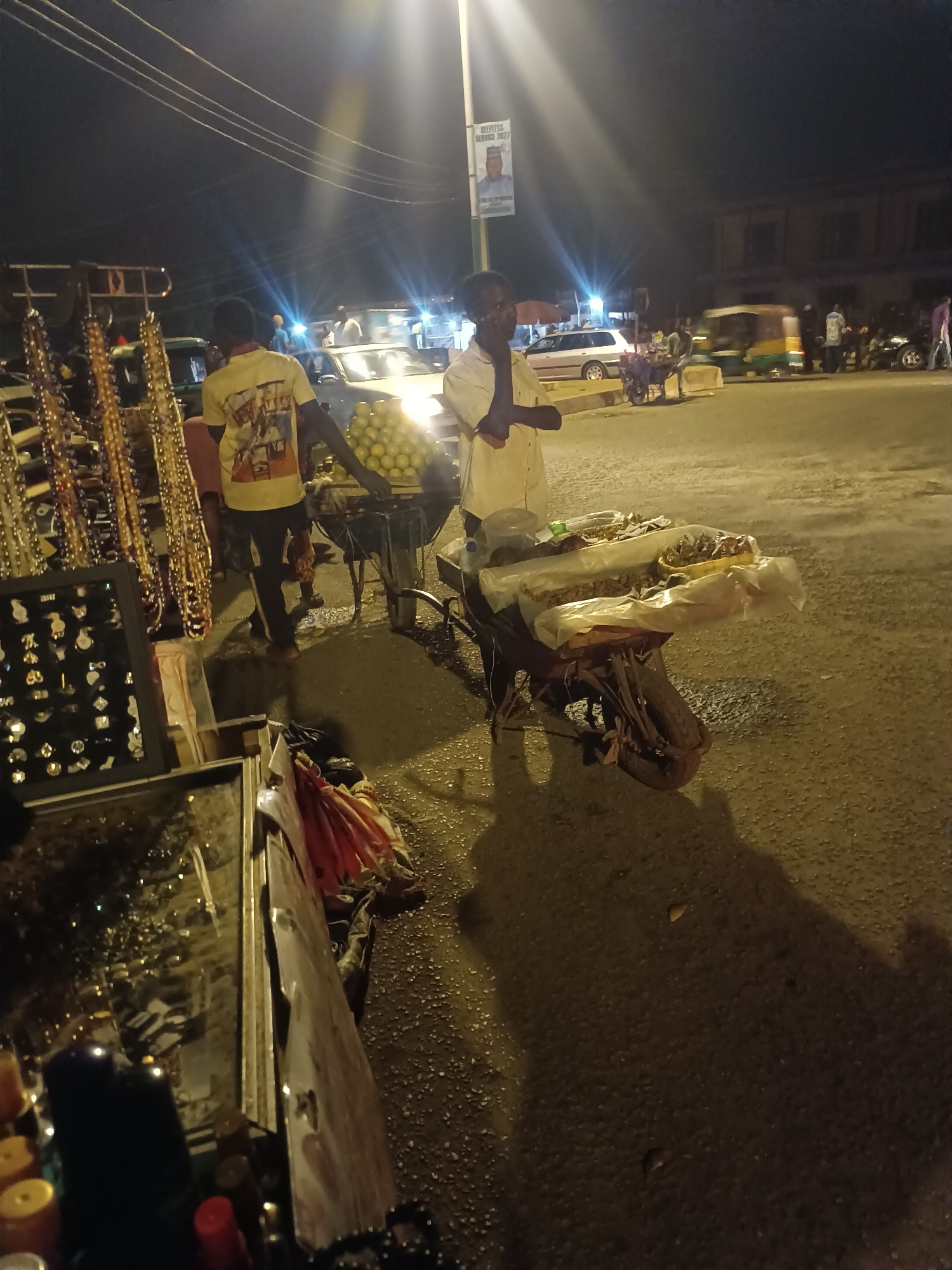 Captured at night in Bukuru, Nigeria, the image presents a street scene dominated by mobile vendors. A male, attired in a light-colored, short-sleeved shirt, stands adjacent to a laden tricycle displaying food items covered in plastic. A second male, wearing a t-shirt with an image on the back and dark trousers, pushes a cart piled high with spherical objects. Another cart, replete with hanging necklaces and displayed earrings, stands closer to the camera on the left. Vehicles, including a yellow taxi and an auto-rickshaw, are visible in the background, along with several pedestrians. The street is illuminated by overhead lighting.
