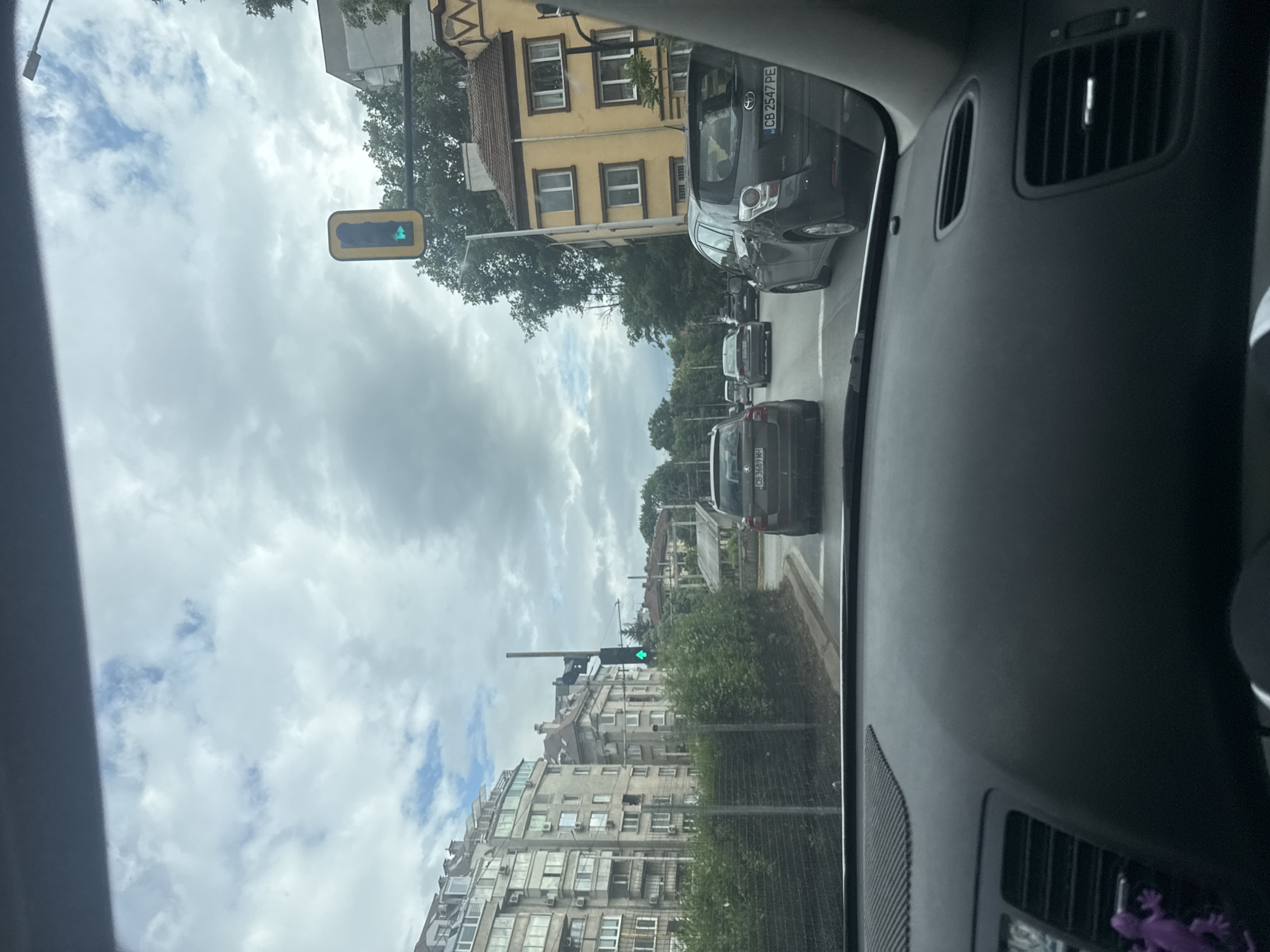 The image is taken from inside a car, looking out the windshield at a street in Sofia, Bulgaria. The traffic light is green, and there are several cars ahead. The buildings on either side of the street are a mix of older, yellow buildings and newer, apartment-style buildings. The sky is cloudy. A purple gecko toy is visible on the dashboard of the car.