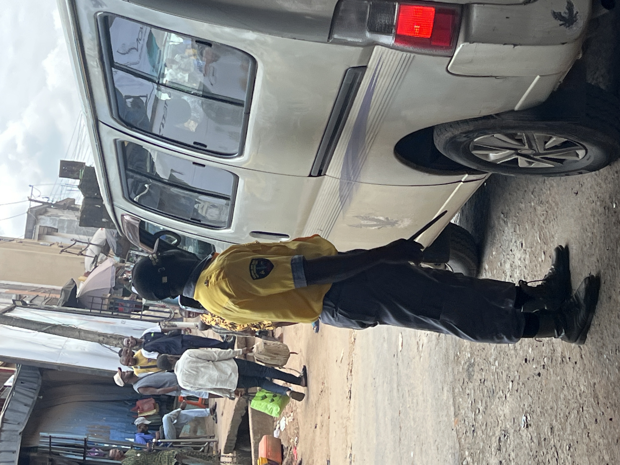 In Nkpor, Nigeria, a Black man wearing a yellow uniform shirt with a dark collar and sleeve trim, a black cap, and dark pants stands next to a silver van. He is facing away from the camera and appears to be holding a baton in his right hand. His black combat boots are laced. The van has tinted windows and a dark stripe with a feathered design along its side. In the background, other individuals are visible. One man, wearing a light-colored shirt and dark pants, carries a green shopping bag and a beige backpack. Other people are seen in the distance, some standing and others seated. The environment appears to be a street scene with buildings and what looks like market stalls in the background. The ground is a dusty, unpaved surface.