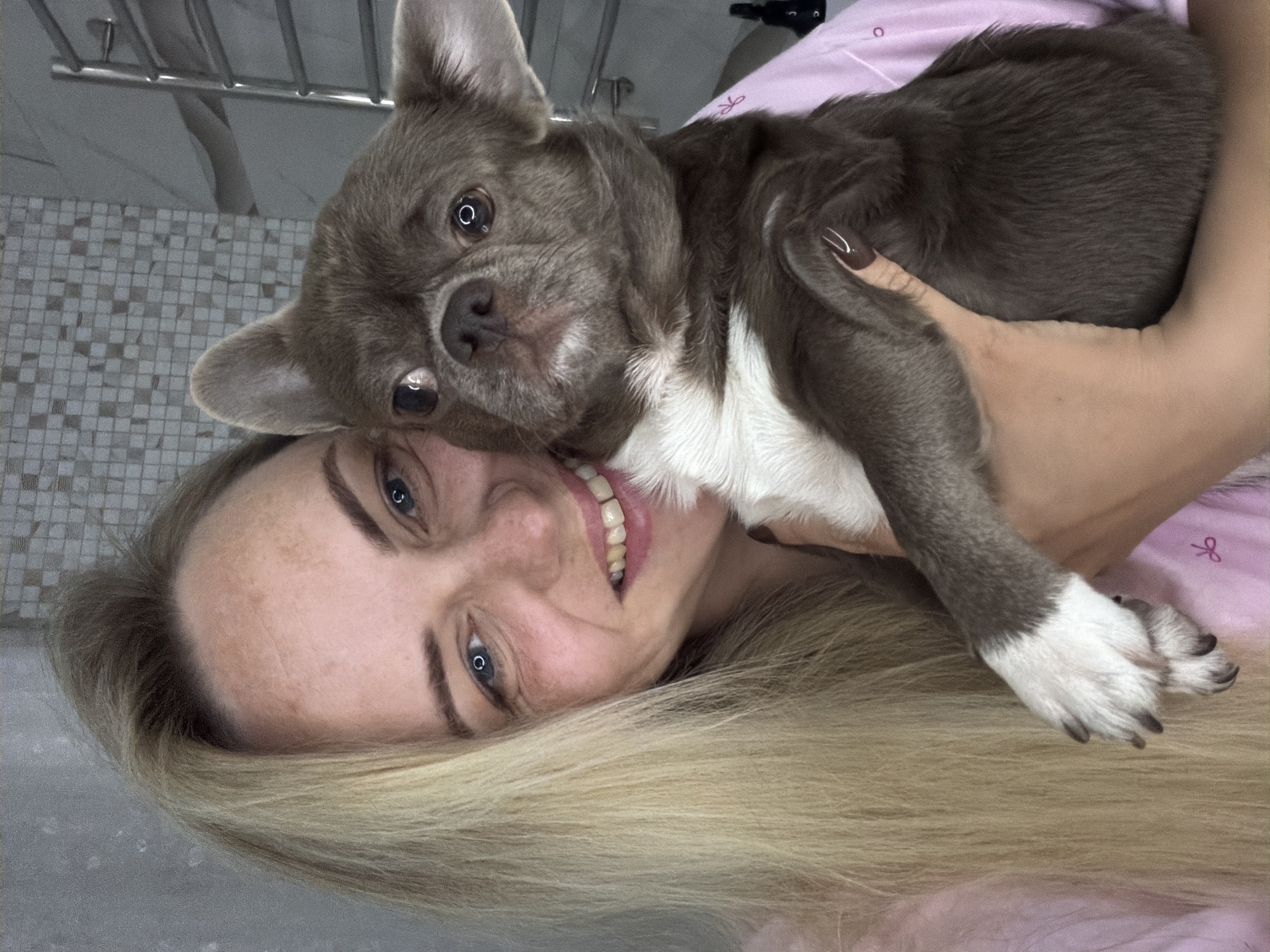 A blonde woman with blue eyes and freckles is holding a small, grey dog with white markings. She is smiling and looking at the camera. The dog is looking slightly away from the camera with its ears perked. They are both wearing pink clothing. The background appears to be a bathroom with tiled walls and a towel rack. The lighting is bright and natural. The scene is likely indoors in a private residence within Votkinsk, Russia.