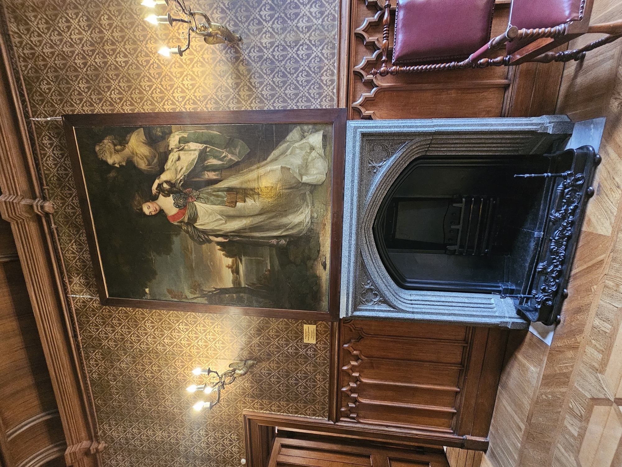 The image shows a room interior. On the wall is a framed painting of a woman in a light-colored dress being attended by another person. The wall has patterned wallpaper. Below the painting is a stone fireplace. To the right of the fireplace are wooden panels and a chair with a maroon cushion. Above the scene, hanging from the ceiling, is a chandelier with multiple light bulbs. The location is likely a building interior, but specific details that would place it within Алупкинский городской совет, Ukraine are not evident from the image itself.
