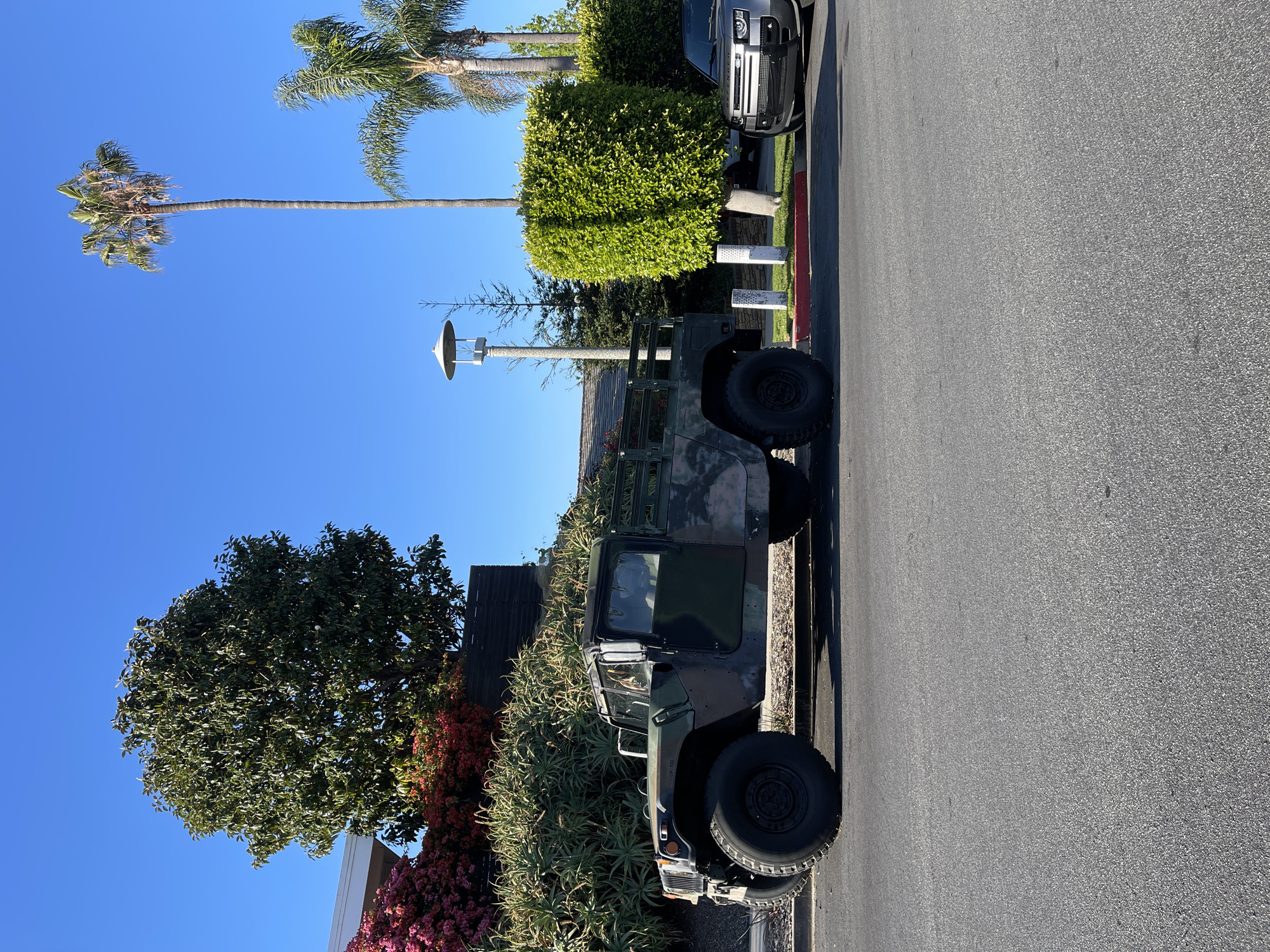 The image depicts a Humvee military vehicle parked on a street in Newport Beach, California. The vehicle is positioned parallel to the curb, near landscaping that includes succulents, trees, and bushes with red and pink flowers. Further down the street, there's a manicured bush and a modern black SUV. Palm trees are visible in the background against a clear blue sky. A street lamp stands nearby, contributing to the residential atmosphere. The scene suggests a sunny day in an affluent residential area.