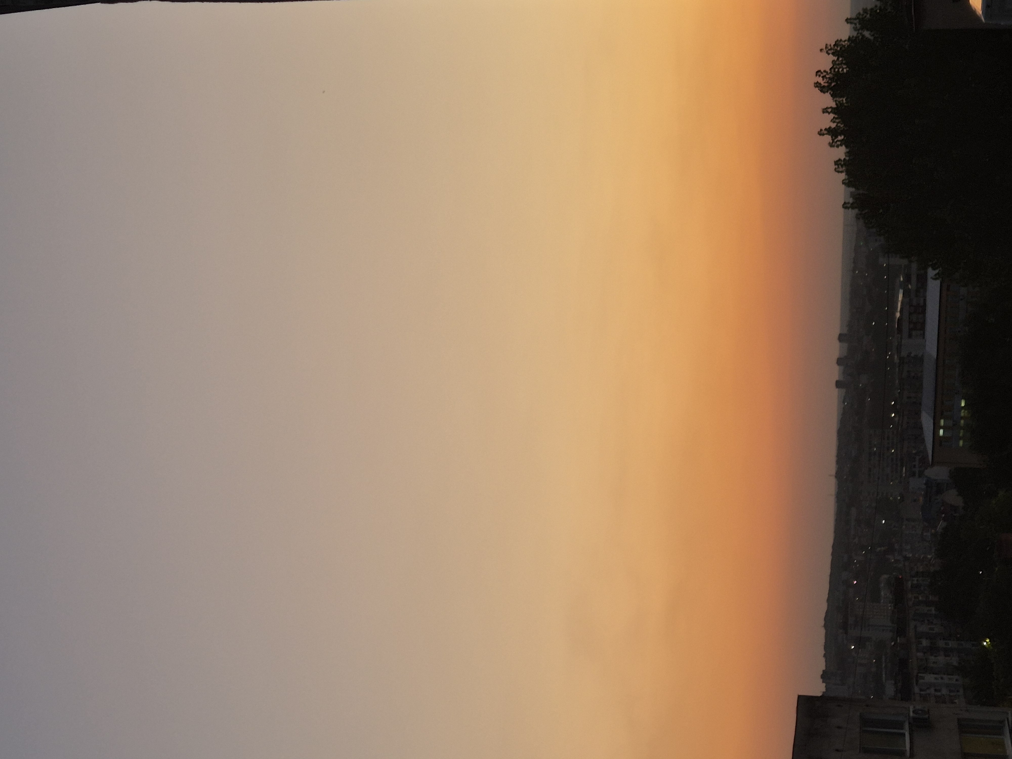 A high-angle, full shot captures a city skyline at dusk. The sky transitions from a light lavender at the top to a warm orange near the horizon. In the foreground, the corner of a concrete building with visible windows and an air conditioning unit juts in from the left. The middle ground is filled with a dense collection of buildings of varying heights, some with lights illuminated. To the right of the frame, a dark, leafy tree dominates the foreground. The overall atmosphere is serene and still.