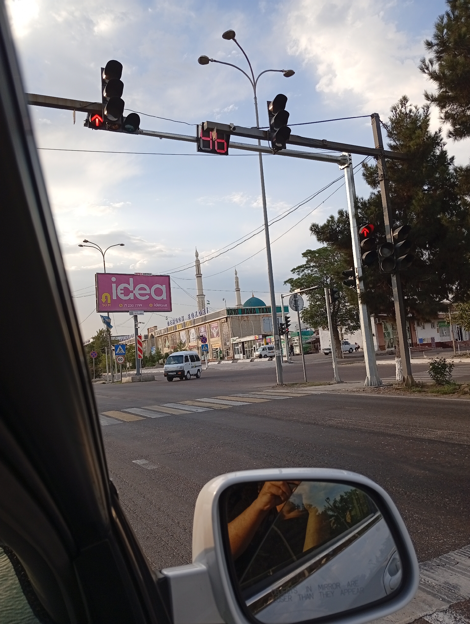 A traffic intersection in Ohangaron, Uzbekistan, is captured from inside a vehicle. A traffic light displays a red arrow pointing upwards. In the background, a white minivan is crossing the intersection, and a building with a domed roof, possibly a mosque, is visible. A large billboard with the word "idea" in pink letters is also prominent. The sky is cloudy, and trees line the side of the road. The reflection in the car's side mirror shows a person's arm and hand.