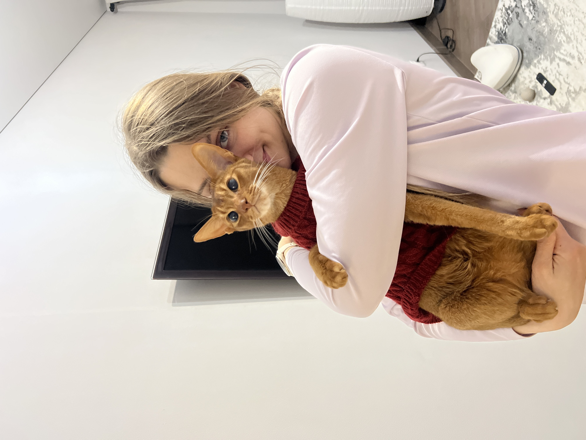 A young woman with blonde hair and fair skin is captured indoors, holding an orange-ginger cat. She wears a light pink long-sleeved top and smiles warmly, gazing directly at the camera. Her arms are wrapped around the cat, holding it affectionately. The cat, likely an Abyssinian breed, is wearing a dark red knitted sweater and also looks towards the camera with a calm and content expression.
The setting appears to be a modern, brightly lit room, possibly a home or apartment, characterized by plain white walls. In the background, a television or monitor is partially visible on the wall behind the woman. To the upper right, there's a glimpse of light-colored furniture, a white object resembling a speaker, and a black remote control on a textured surface. The lighting is soft and even, suggesting daytime. No specific location cues beyond an indoor environment are discernible, though the image is contextualized as being taken in Kazan, Russia. The overall mood is one of warmth, affection, and comfort between the woman and her pet. No visible text is present in the image.