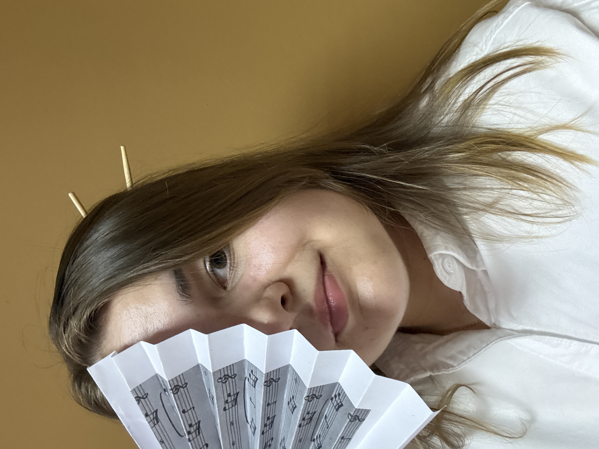 A woman with light brown hair is holding a white paper fan with printed musical notes in front of her face. She is wearing a white collared shirt. Two wooden chopsticks are stuck in her hair. The background is a plain, warm yellow-orange color. The image appears to be taken indoors. There is no indication of the location being Chaikovsky, Russia.