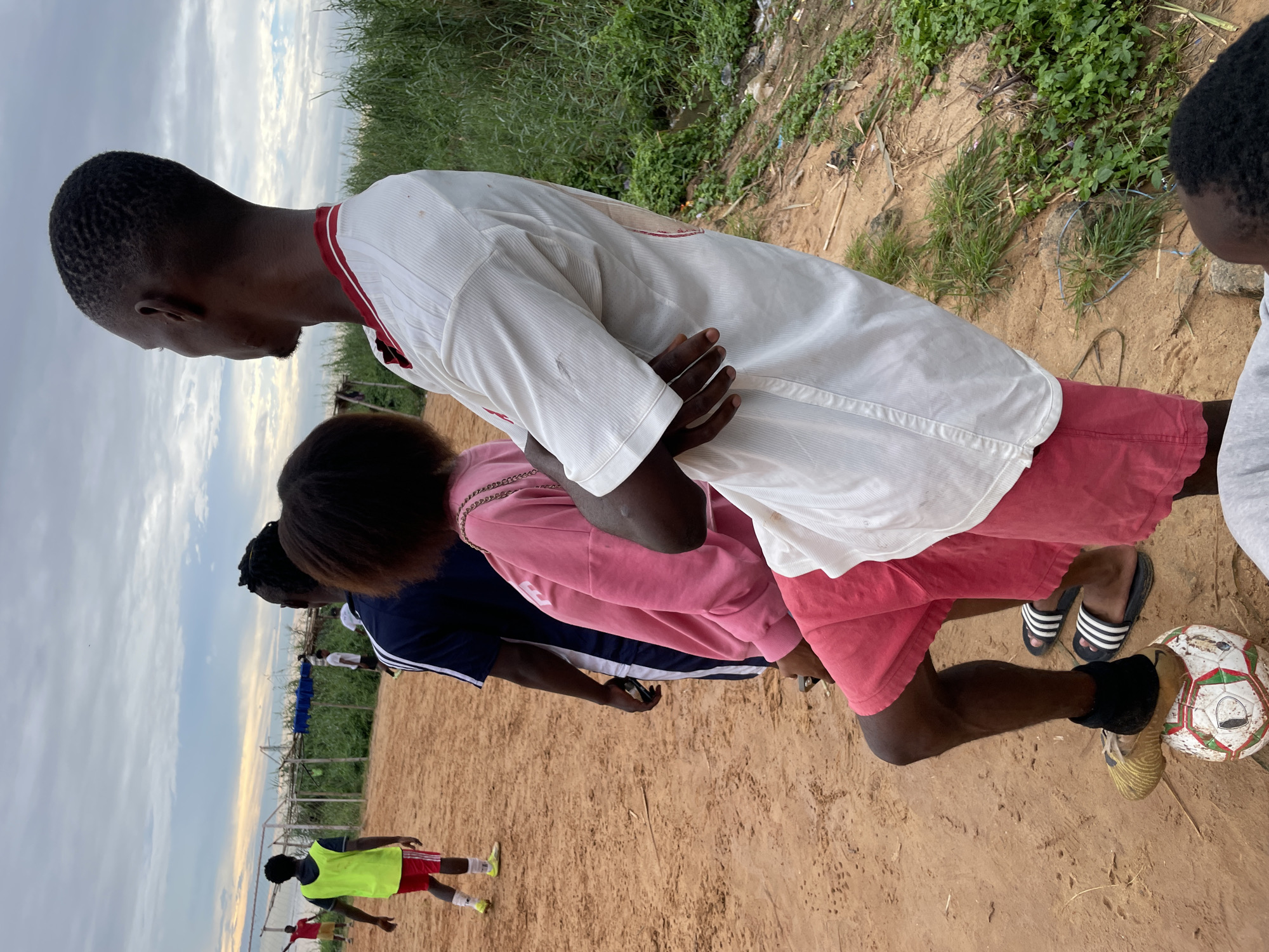 A group of people are gathered on a dirt field, possibly for a soccer game, with a fence and vegetation in the background. A young man in the foreground, wearing a white t-shirt and red shorts, stands with his arms crossed, his right foot resting on a soccer ball. To his left, a young woman in a pink sweatshirt and matching shorts is also standing. Behind her, another person in a dark blue and white striped shirt is visible. Further in the background, several other individuals are scattered on the field, some appearing to be playing soccer. The sky is overcast with clouds. The scene appears to be an outdoor recreational area, possibly in Ado, Nigeria.