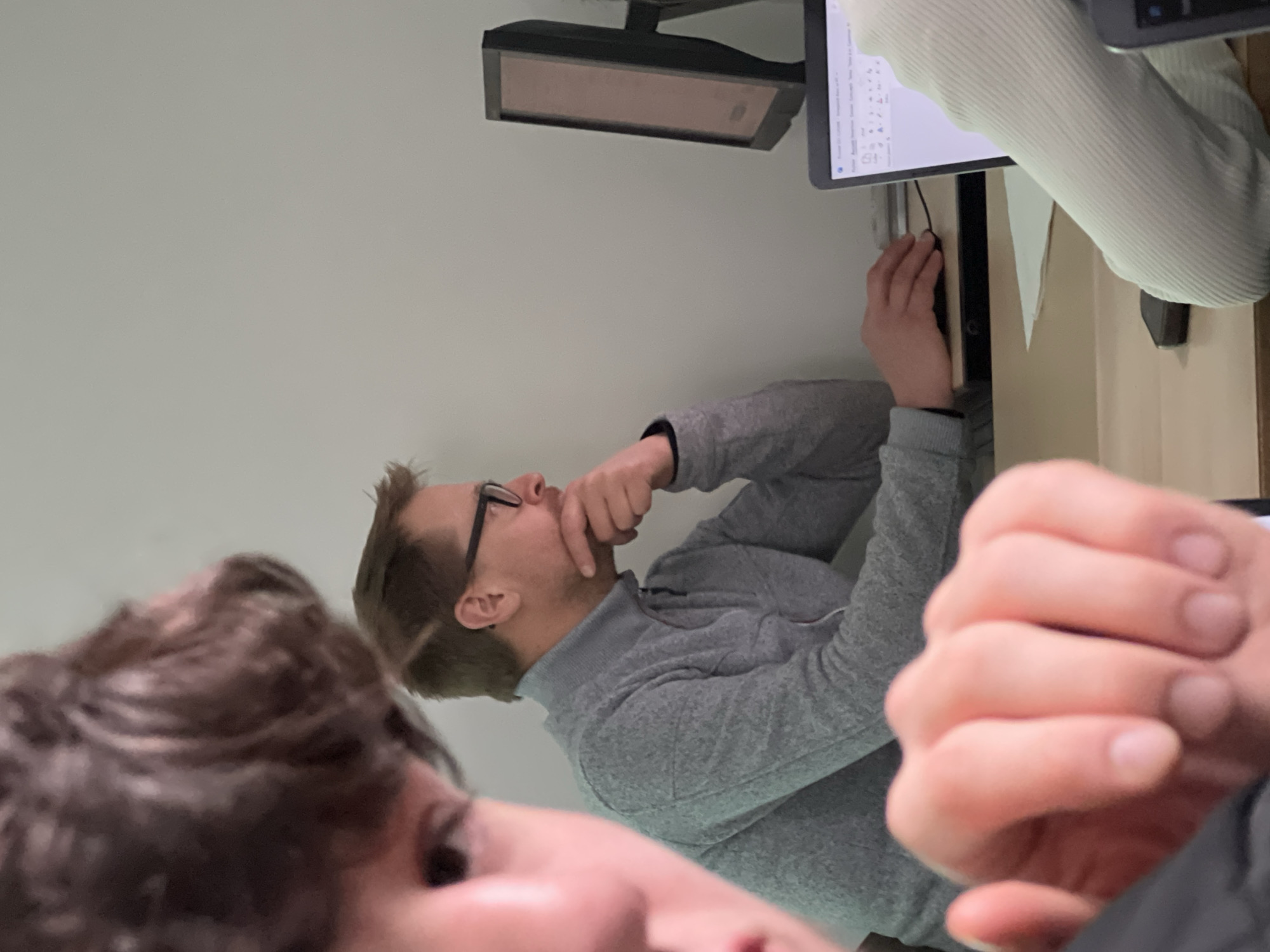 The image captures an indoor scene. A man with glasses, wearing a gray sweater, is looking at a computer screen with his hand near his mouth as if in thought. Another person, with curly brown hair, is partially visible in the foreground. A second person, wearing a light-colored sweater, is operating a computer mouse. Two computer monitors and a desk are also visible in the frame. There is no information given to determine that this image was captured in the city of Schiltigheim, France.