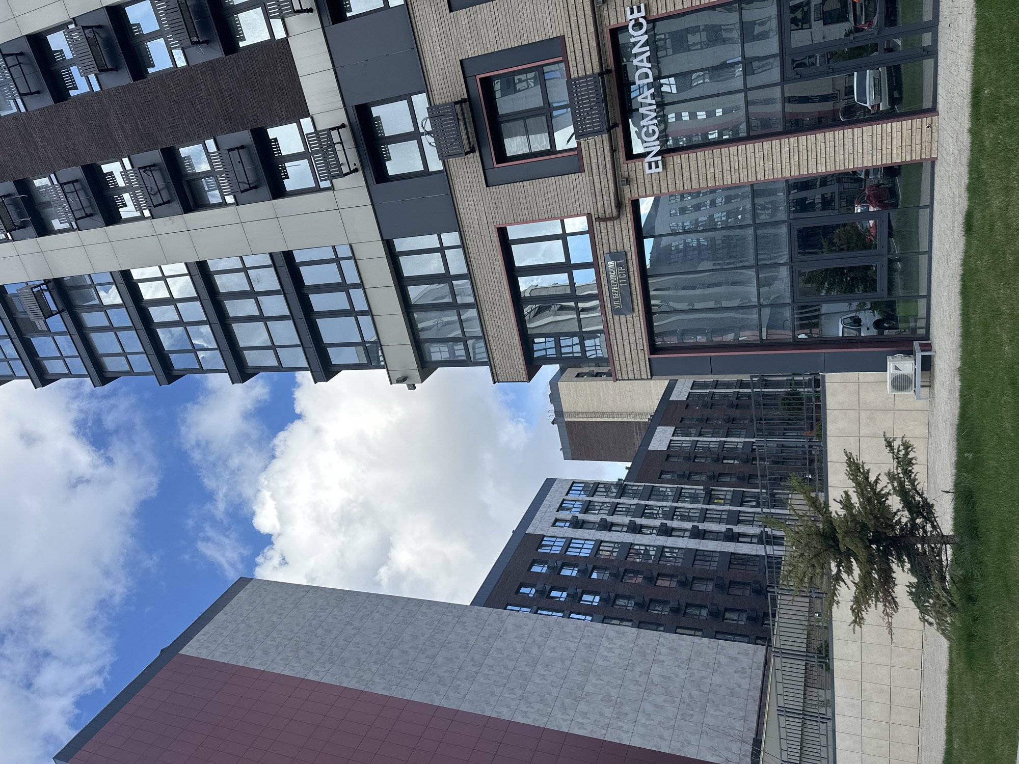 The image shows a street view in Rostov-on-Don, Russia. A multi-story building with the signage "Enigma Dance" is visible, along with other modern apartment buildings with numerous windows reflecting the sky. The building's facade is a combination of brick, gray, and glass elements. Street signage with Cyrillic text is also present. In the foreground, there is greenery, a tree, and a paved walkway. The sky has a mix of blue and cloudy patches.