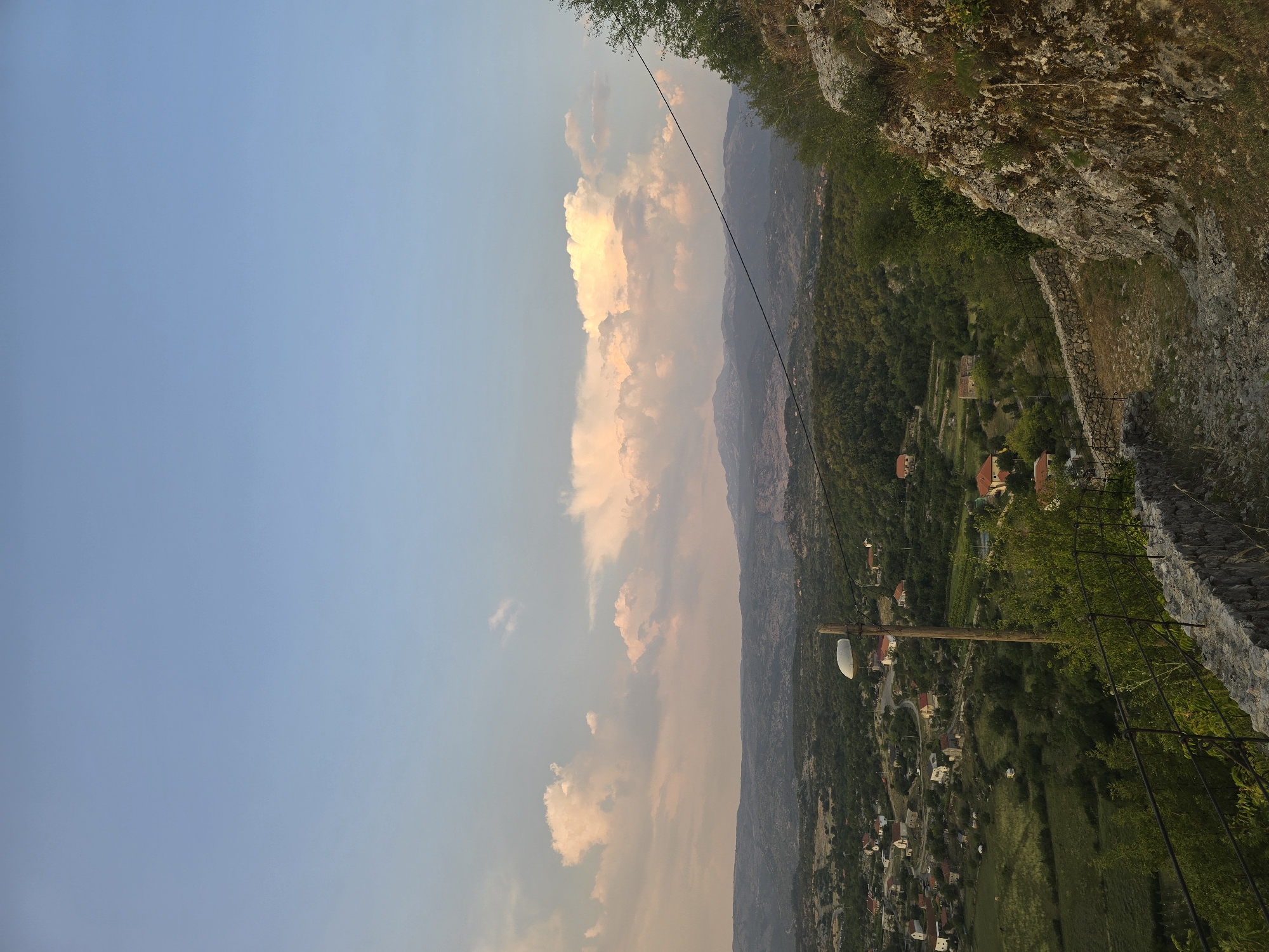 The image shows a landscape view of Medun, Montenegro, featuring rolling green hills dotted with houses, a stone-walled pathway, and a clear blue sky with scattered clouds. A wooden pole with a street lamp stands prominently in the foreground, alongside a wire strung across the scene. The rocky terrain on the right side of the image suggests a natural, rugged environment. The lighting indicates it is likely late afternoon or early evening.