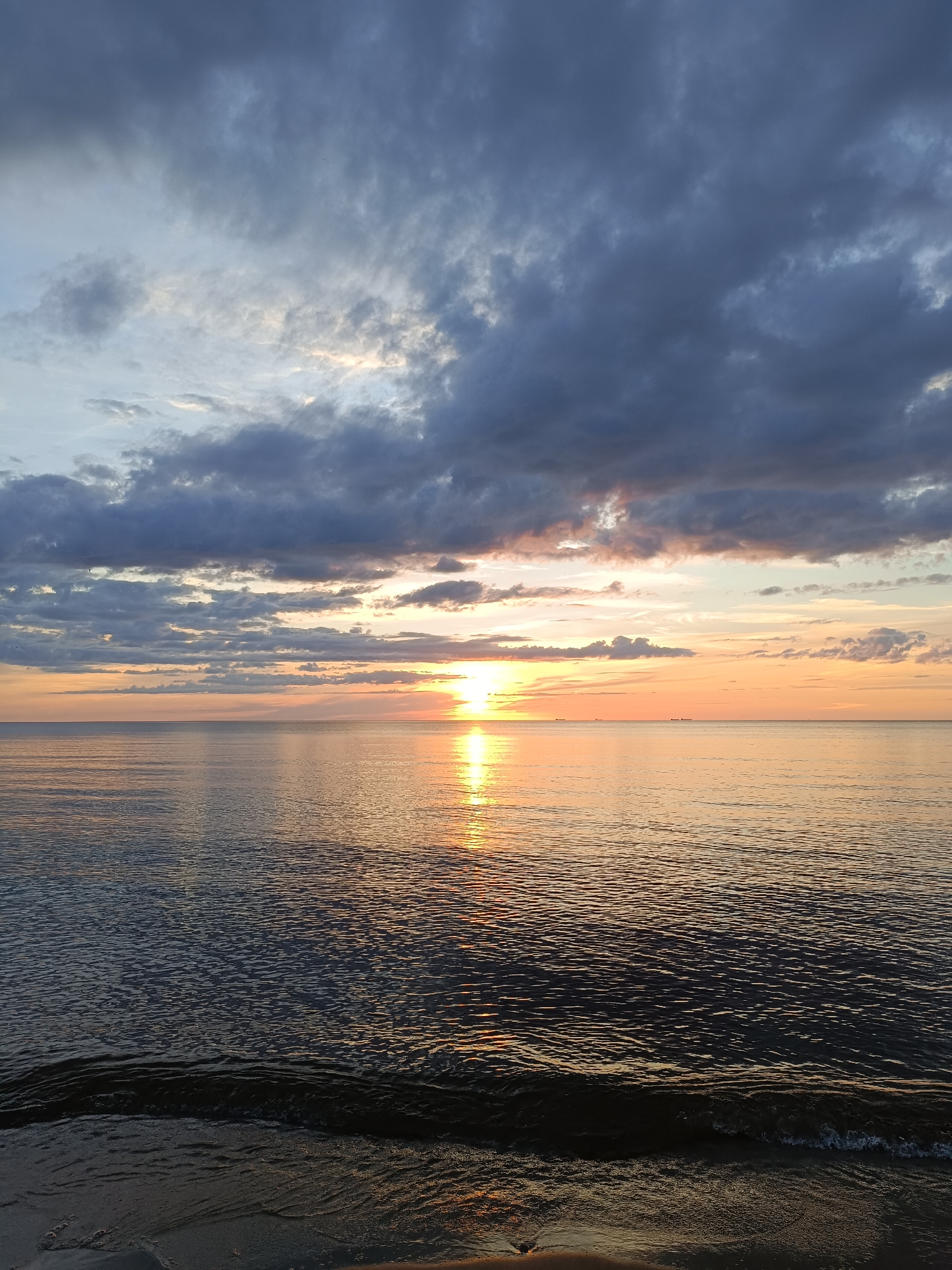 A serene sunset paints the sky with hues of orange and yellow, reflected on the calm sea. Dark, textured clouds dominate the upper portion of the image, contrasting with the lighter, brighter sky near the horizon. The sun, a brilliant orb, is positioned slightly left of center, casting a shimmering path of light across the water. A gentle wave laps at a dark, sandy shore in the foreground. The scene appears to be a natural landscape, likely a beach at dusk. There are no people, animals, or man-made structures other than distant, almost imperceptible, shapes that might be ships on the horizon. The overall mood is peaceful and tranquil.