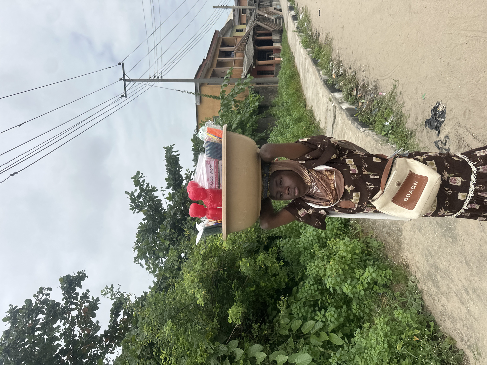 A young woman stands on a dirt road in Badagry, Nigeria, with a large bowl of merchandise balanced on her head. The bowl contains various items, including red plastic objects, packaged hair extensions, and other assorted goods. She is wearing a patterned brown dress and a headscarf, with a cream-colored bag slung over her shoulder. To the left of the woman, lush green vegetation lines the road. In the background, buildings with balconies and weathered architecture are visible. Overhead, power lines crisscross the sky, which is overcast with clouds. The scene depicts a typical street vendor in a Nigerian city.