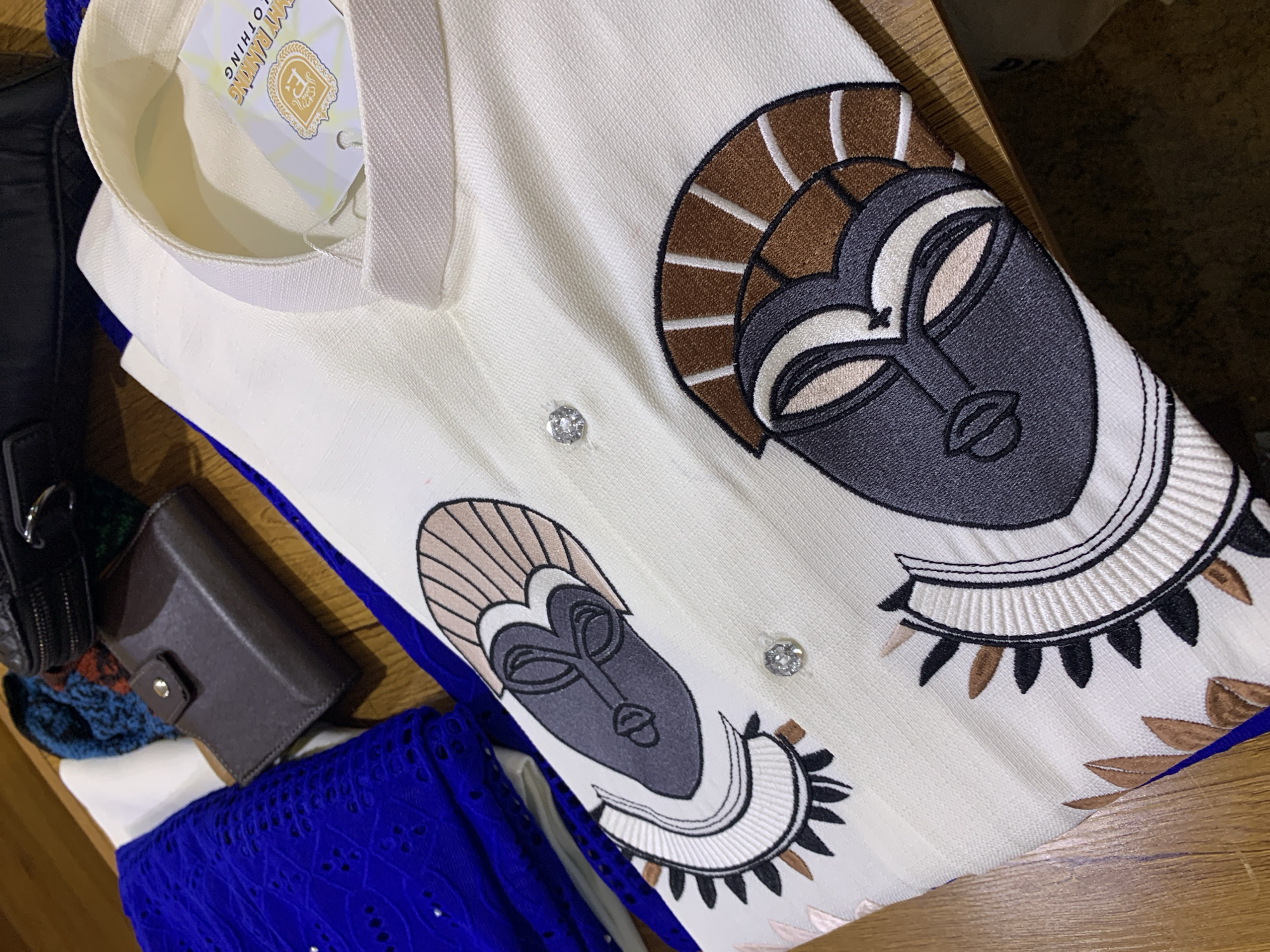 This image is a close-up, overhead view of a cream-colored garment, likely a traditional Nigerian top, laid out on a wooden surface. The garment features two prominent embroidered depictions of tribal masks in shades of grey, white, and brown, with a distinct circular pattern resembling an eye below each mask. Two small, clear rhinestones are attached to the fabric. A white tag with gold lettering, reading "MY RANKING CLOTHING," is partially visible near the collar. To the left of the garment, a folded piece of vibrant blue fabric with an eyelet pattern is visible, along with a small brown leather wallet. In the background, a black zippered pouch and a knitted item with blue, red, and dark green threads are present. The scene suggests a display or preparation of clothing, possibly in a home or market setting within Gura Top, Nigeria.