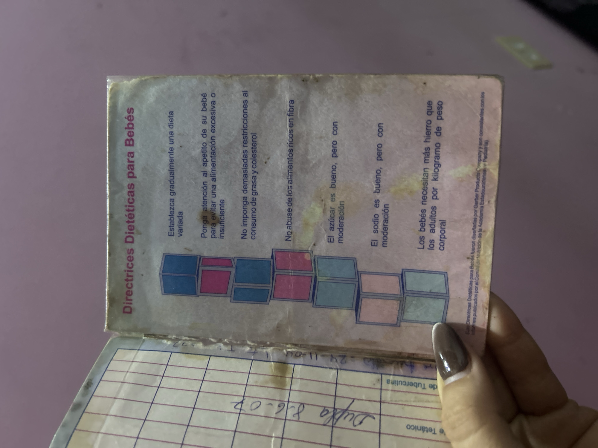 Here is a detailed description of the image:
The image shows a close-up of a partially open booklet titled "Directrices Dietéticas para Bebés" (Dietary Guidelines for Babies). The booklet provides nutritional advice for infants, covering topics such as establishing a varied diet, paying attention to appetite, avoiding excessive restrictions on fat and cholesterol, and moderating sugar and sodium intake. It also states that babies need more iron per kilogram of body weight than adults.
Dates "24-11-04" and "8-6-07" are written on the open part of the booklet along with "Dupla".
A person's hand with a nail is visible.
The booklet appears aged and worn.
There is no information available to determine the image was taken in San Juan de Los Morros, Venezuela.